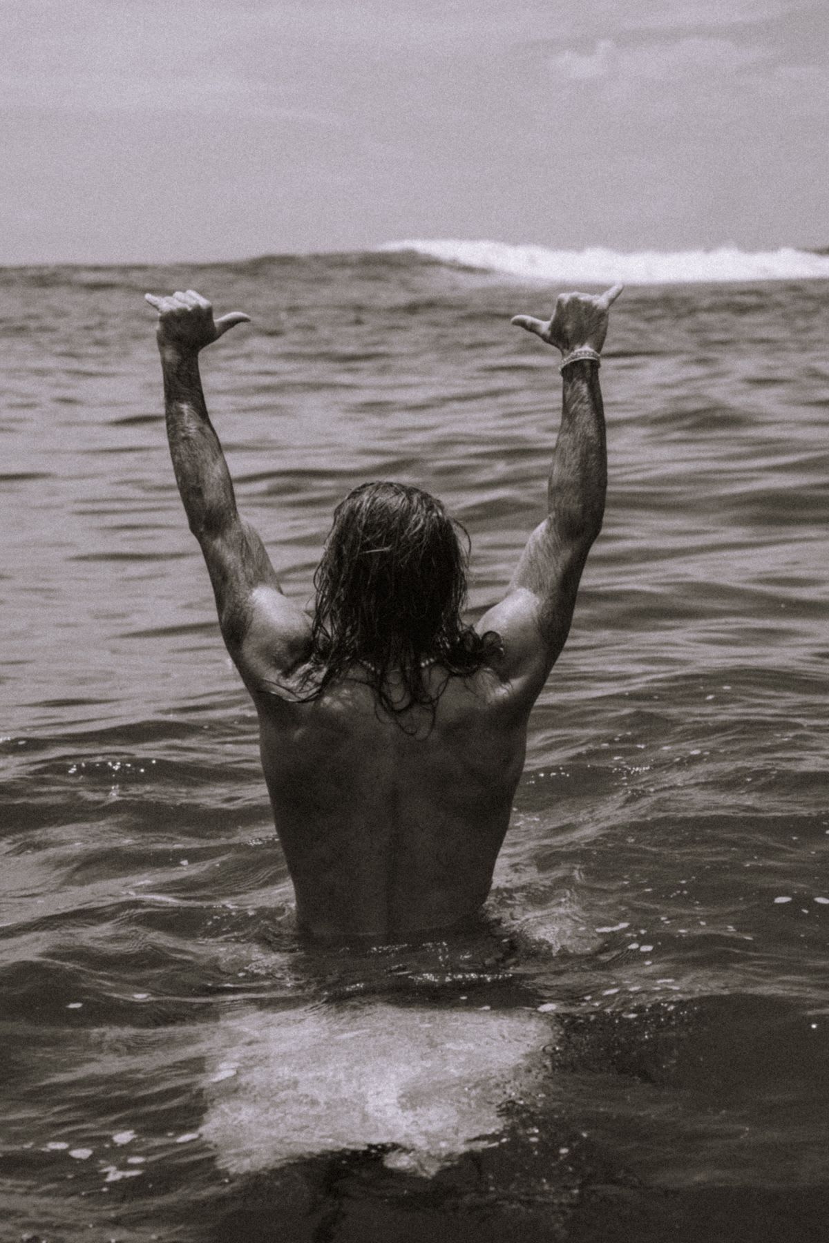 A black and white photograph shows the back of a long-haired man standing in the ocean making a double shaka sign towards a wave.