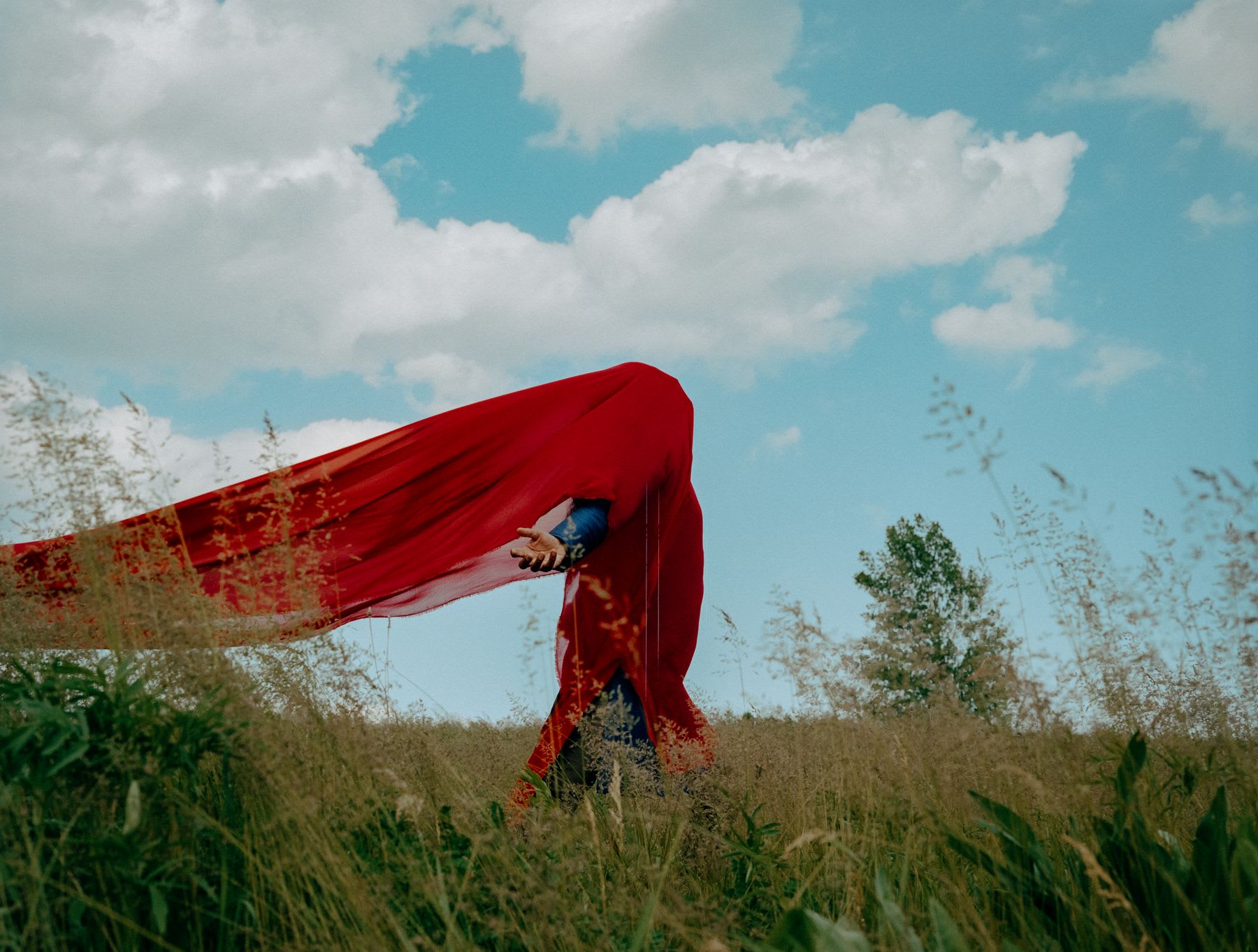 A person in a grassy field is draped in a long red fabric billowing in the breeze under a bright blue sky with scattered clouds.