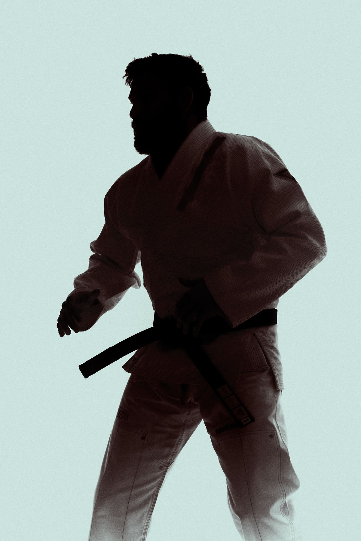 Man in martial arts kimono and black belt adopts a ready stance against a light background.