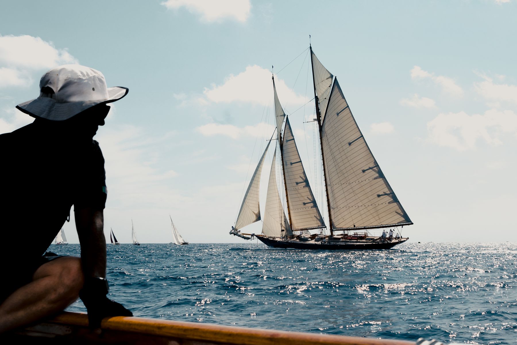 A silhouetted person in a white sun hat watches a classic tall ship with full sails on the sparkling ocean during a regatta.