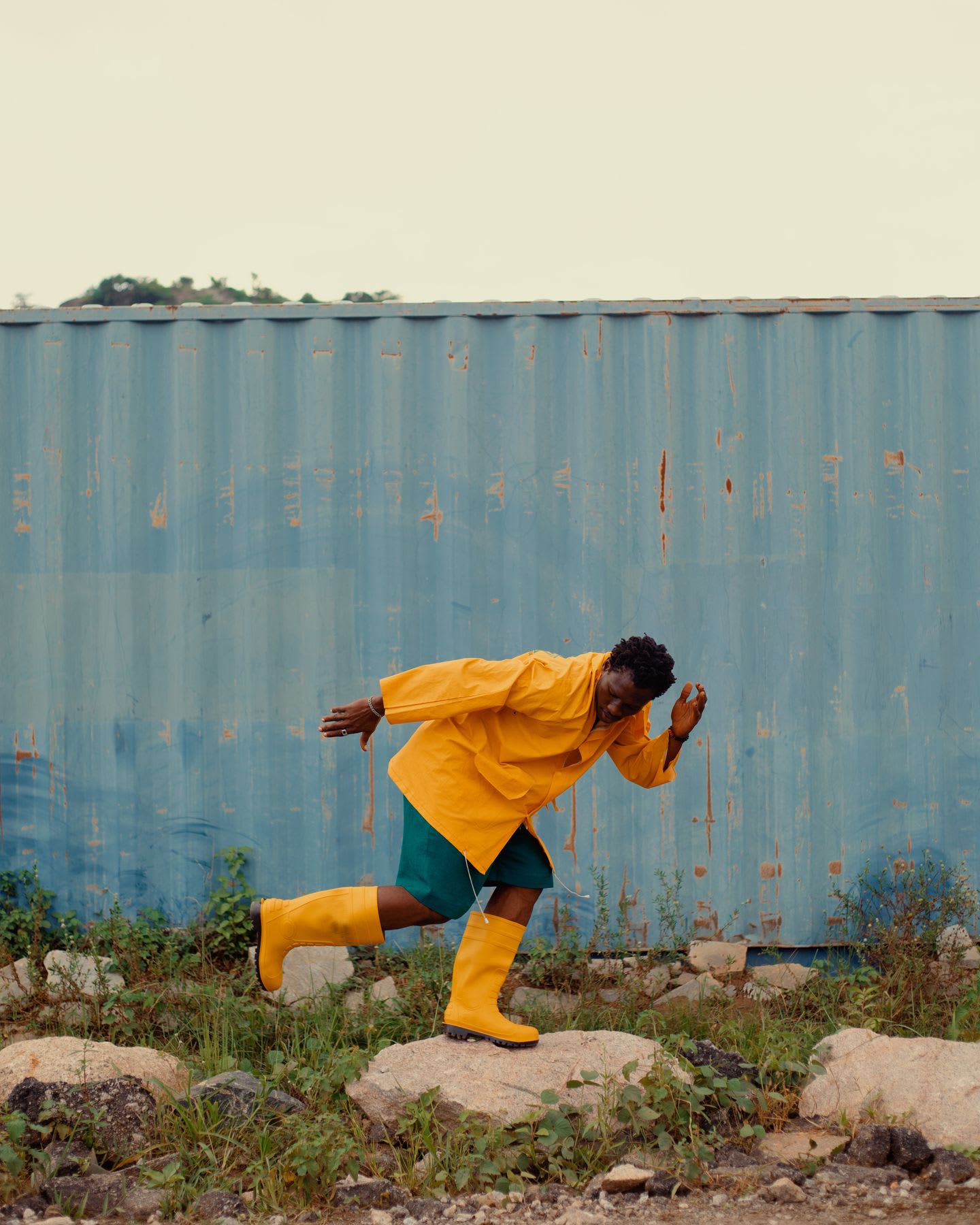 A person in a bright yellow raincoat and matching boots runs across rocks in front of a large blue container.
