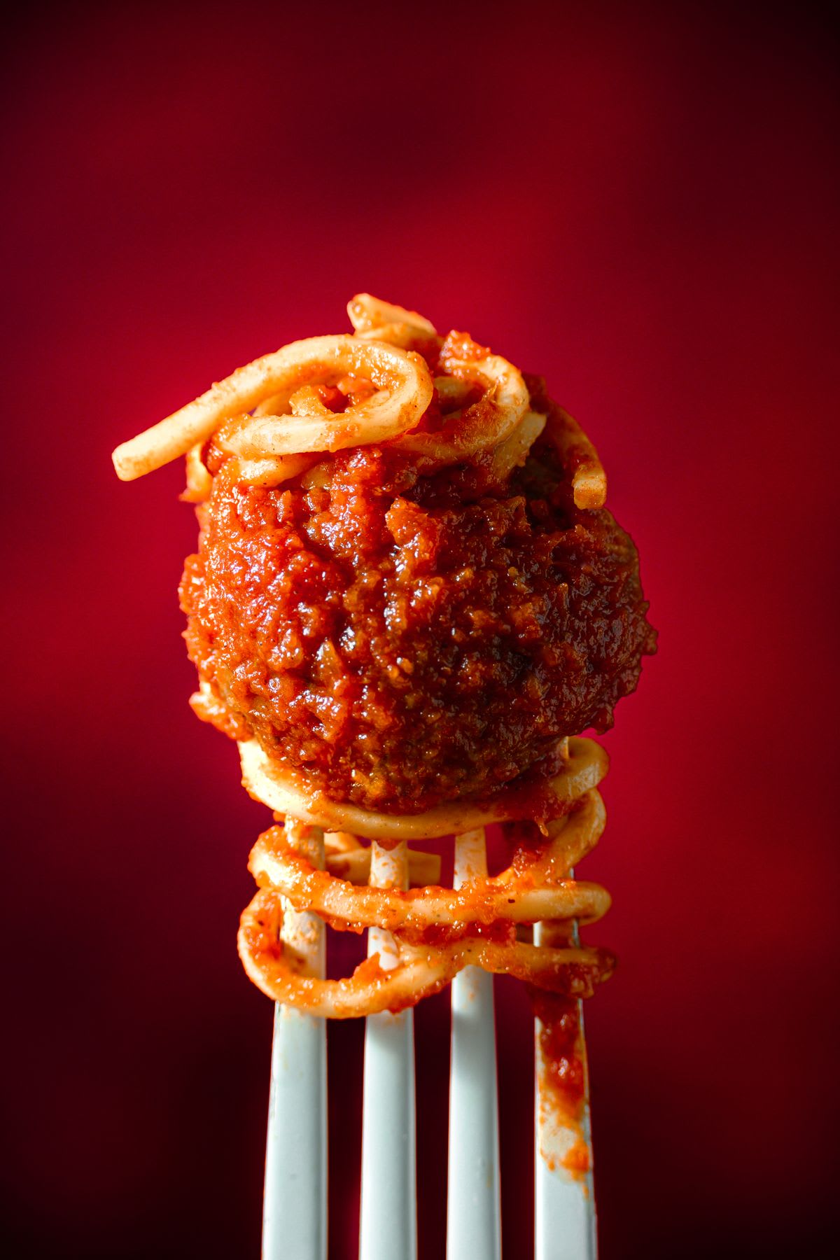 A close-up of a round, brown, and glistening meatball and spaghetti on a metal fork.