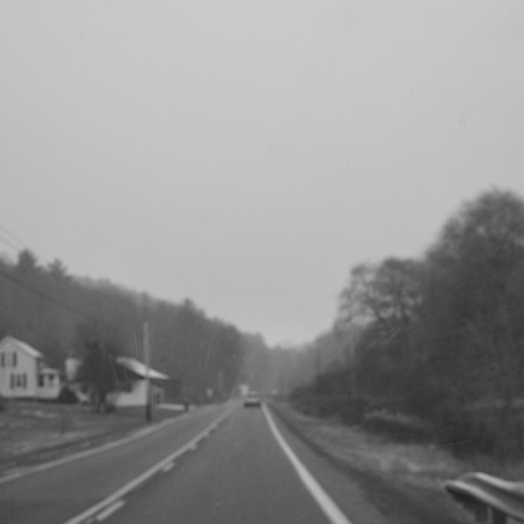 A car drives along a rural highway next to a white, two-story house.