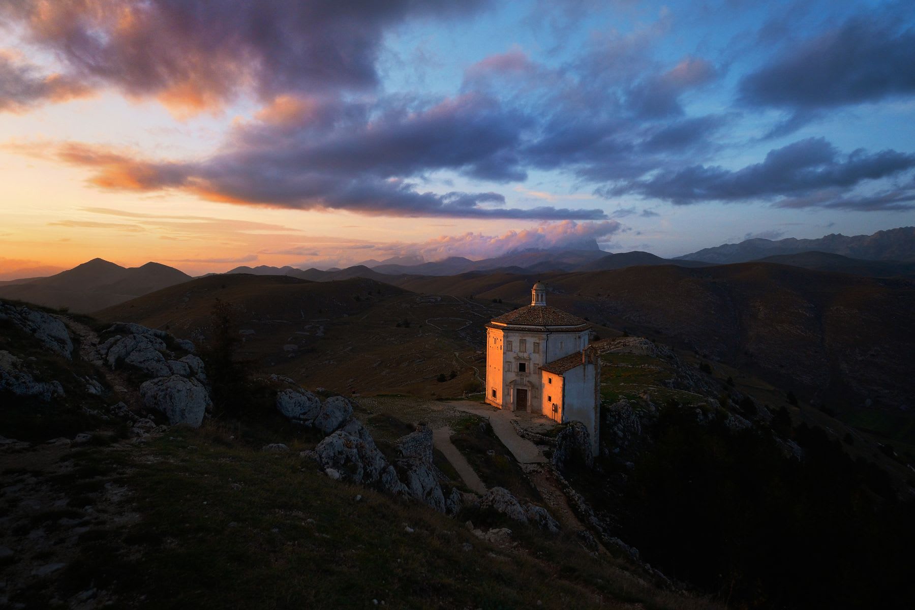 A solitary church, bathed in the warm glow of sunset, perches atop a hill overlooking a vast landscape of rolling hills and distant mountains.