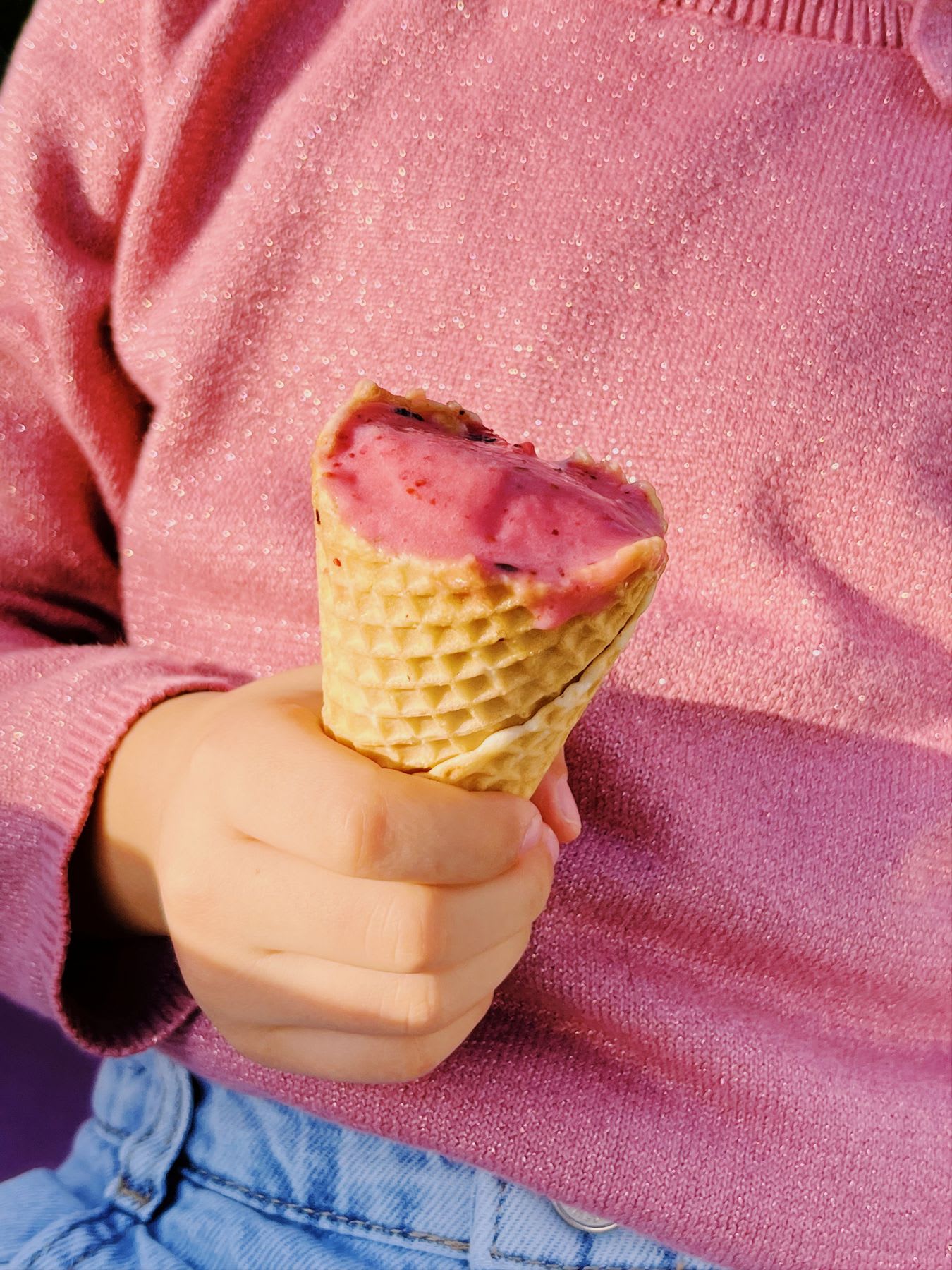 A child in a sparkly pink sweater holds a partially eaten pink ice cream in a waffle cone on a sunny day.