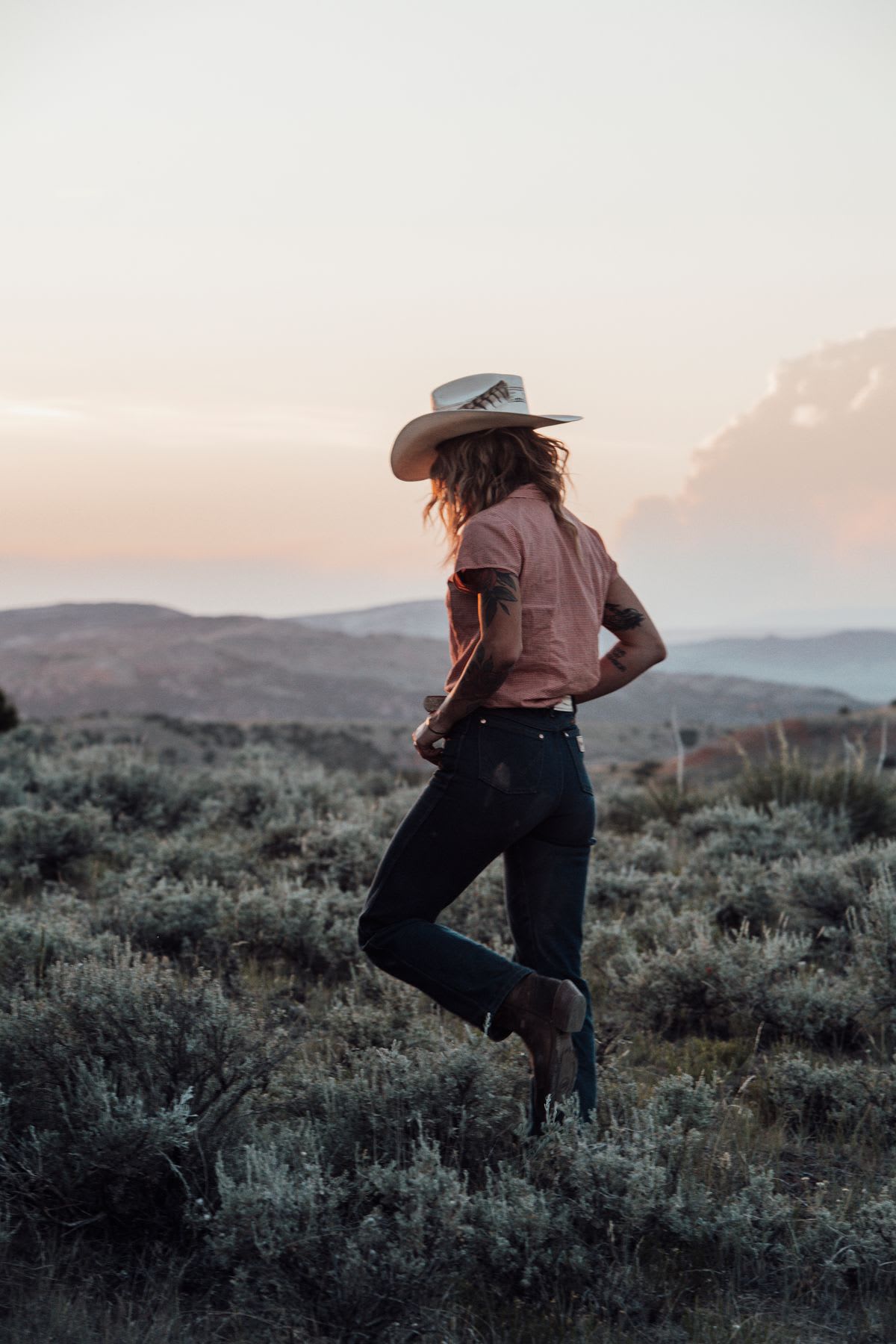 A person in a cowboy hat contemplating the landscape at sunset in an open field.