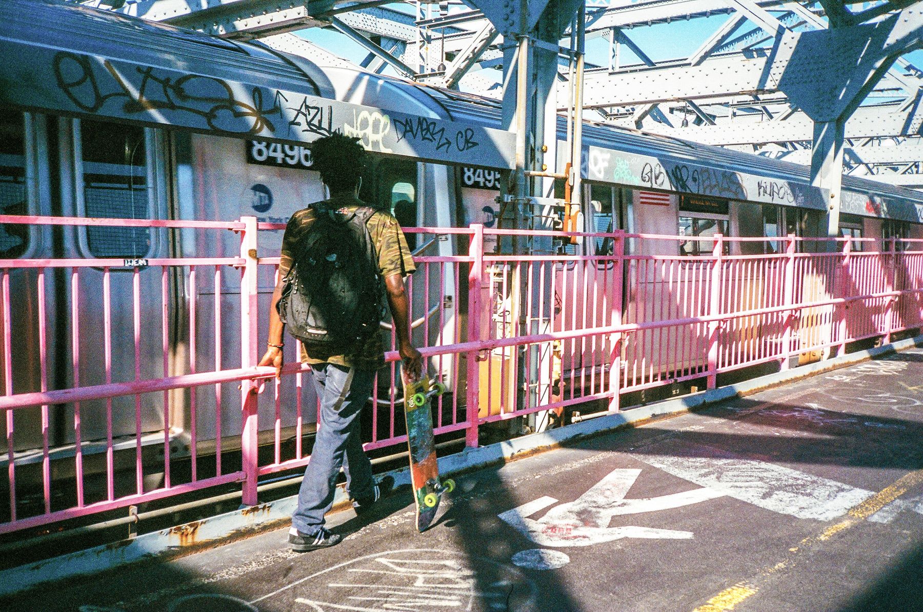 Man carries a skateboard in one hand and holds a pink railing next to the metro train with the other.
