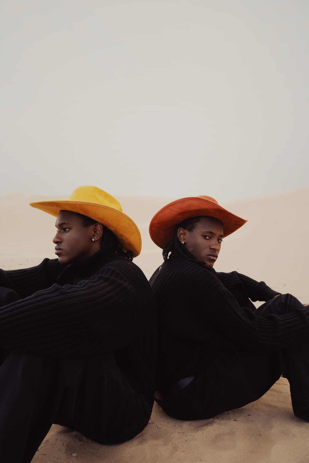Two men sitting back to back in the desert wearing colorful cowboy hats and black clothes.