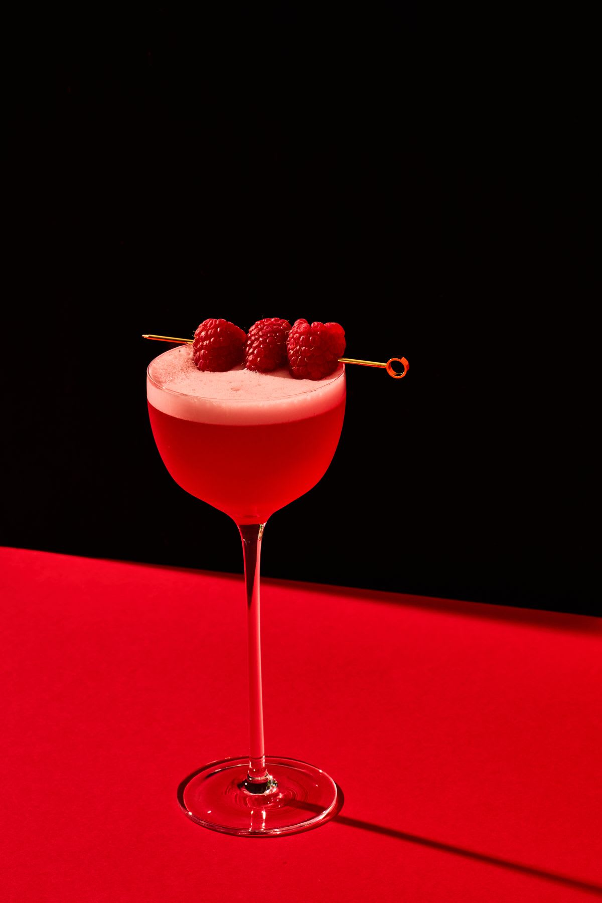 A vibrant red foamy cocktail in a stemmed glass is garnished with fresh raspberries and set on a red table against a black background.