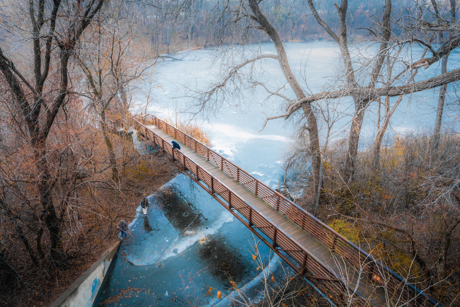 Aerial view of a footbridge across a river in the forest as two people stand in the water along the coast