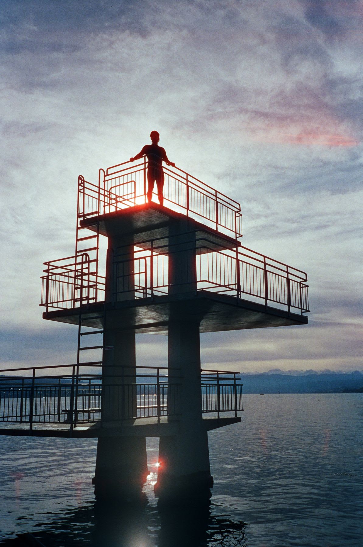 Person standing on top of a diving platform over the water at sunset.