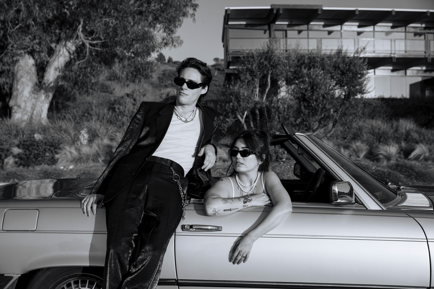 A stylish young man and woman in sunglasses pose with a classic convertible car in this black and white photograph.