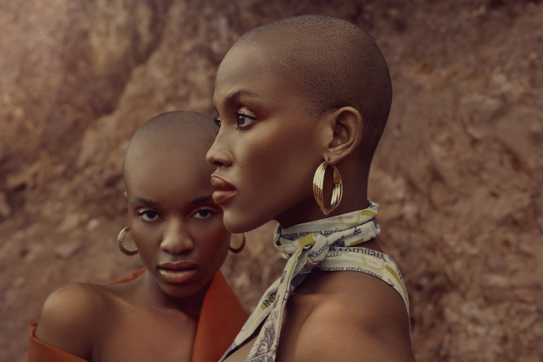 Close-up of two women posing in dresses and gold earrings in a natural setting.