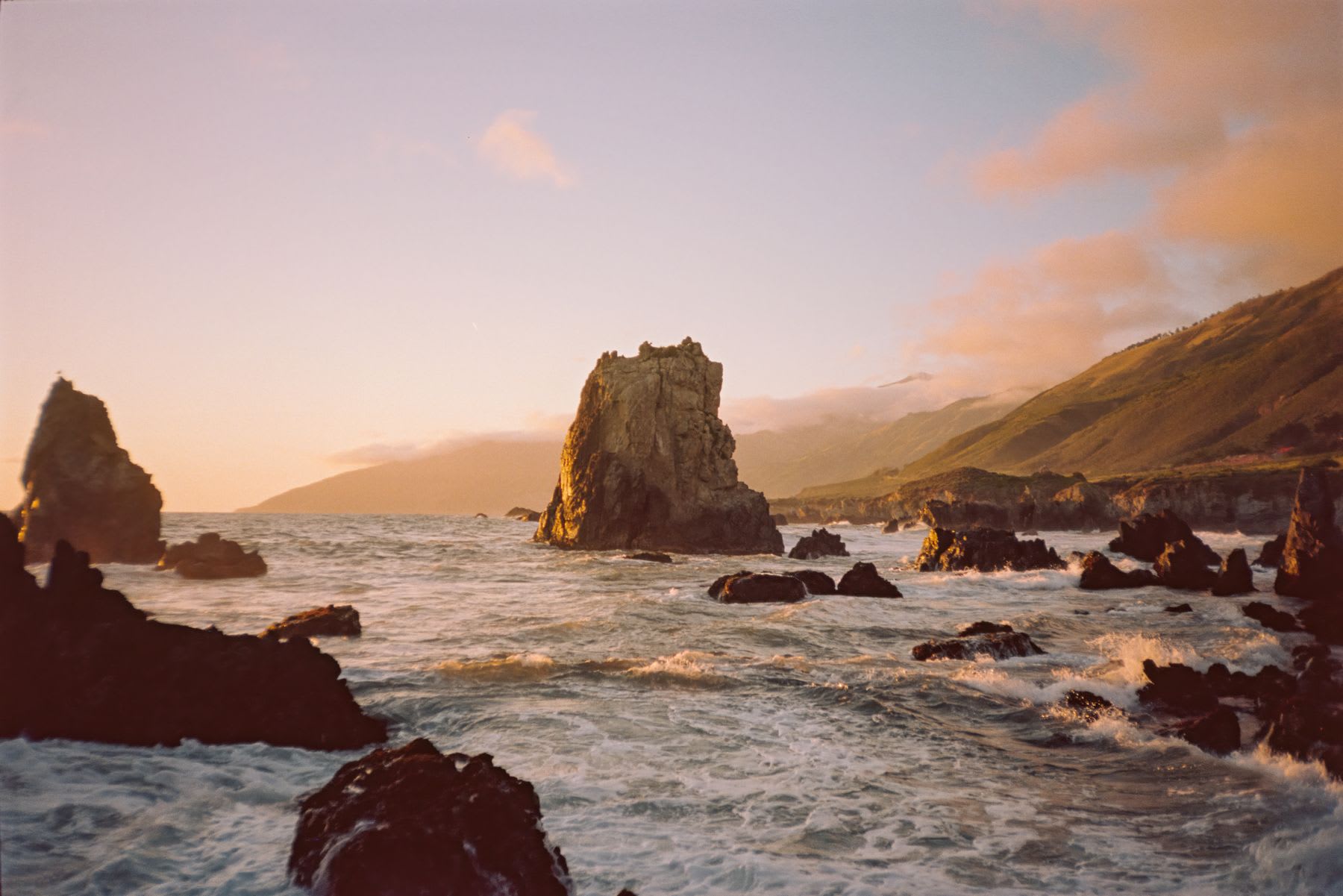 The warm light of dusk illuminates the rugged, rocky coastline as waves crash into the shore.