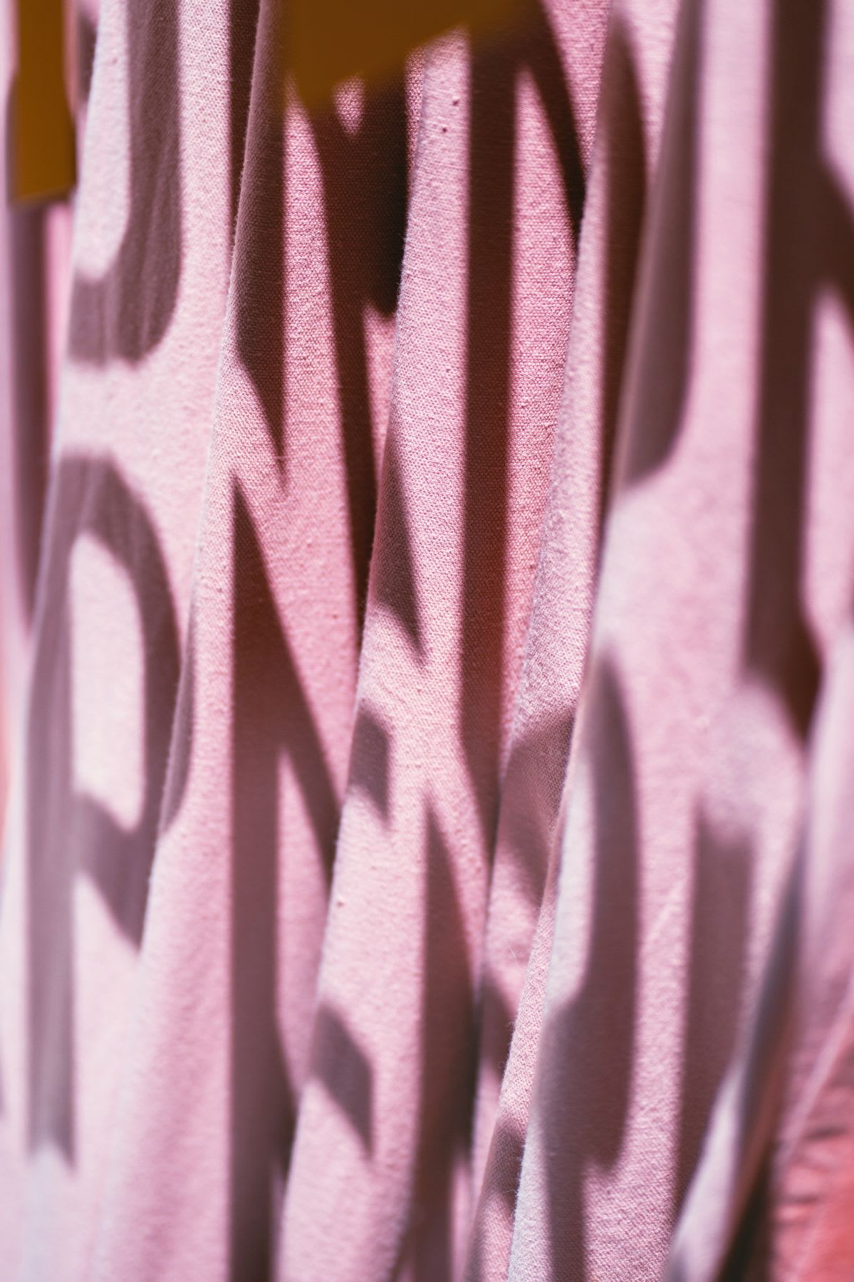Shadows of letters are projected onto pink fabric, creating a graphic and abstract effect.