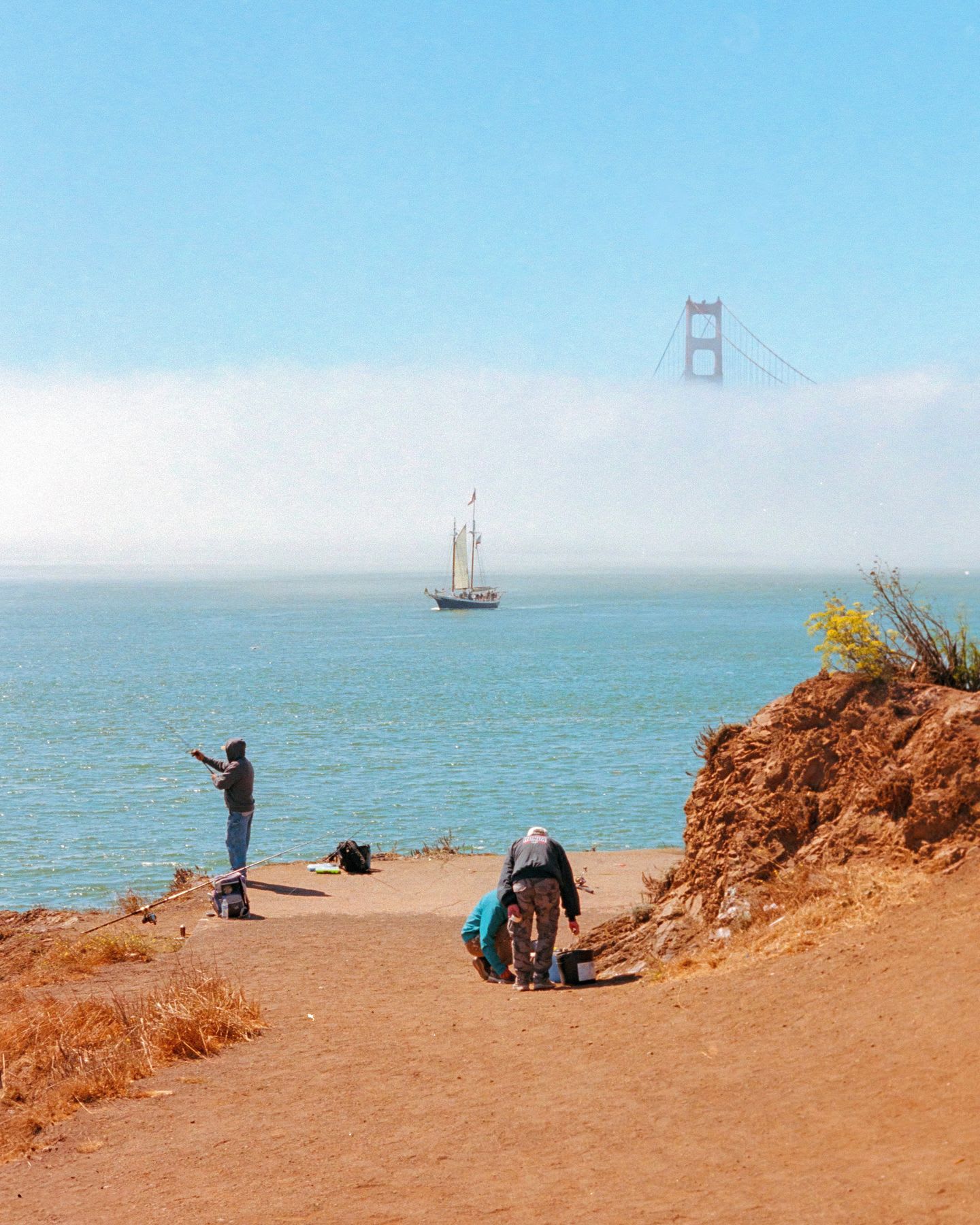 One person in a green coat fishes, while two others, one in a black jacket and one in a blue sweater, pick up items from the reddish-brown shore, with a sailboat on the water and the Golden Gate Bridge partially obscured by fog in the distance.