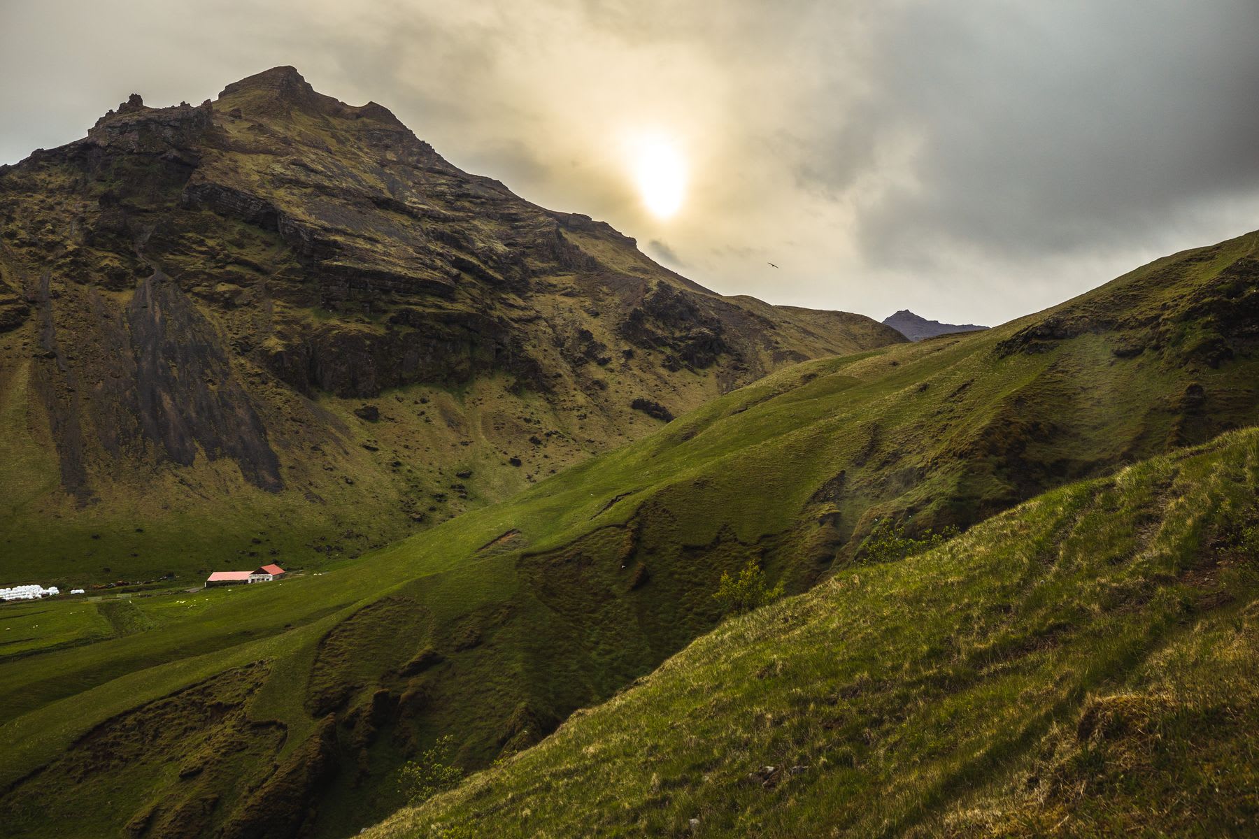 Sunlit mountainside with green slopes and a solitary red-roofed house nestled in a valley, under a cloudy sky.