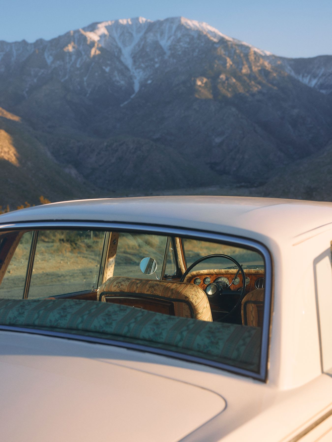 The interior of a vintage car is visible through the rear window, next to a mountain range under a clear sky.