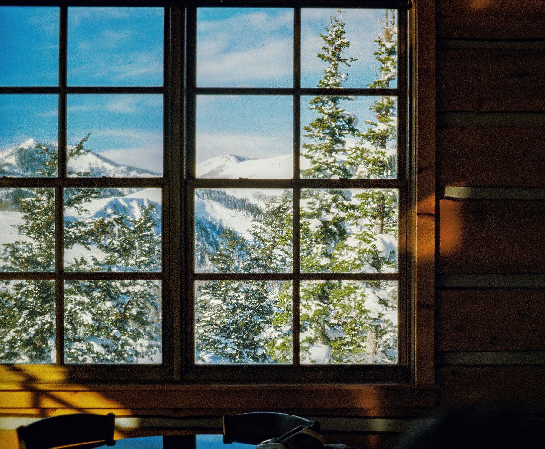 A sunlit wooden cabin interior frames a picturesque view of snow-covered mountains and evergreen trees through a large, multi-paned window.