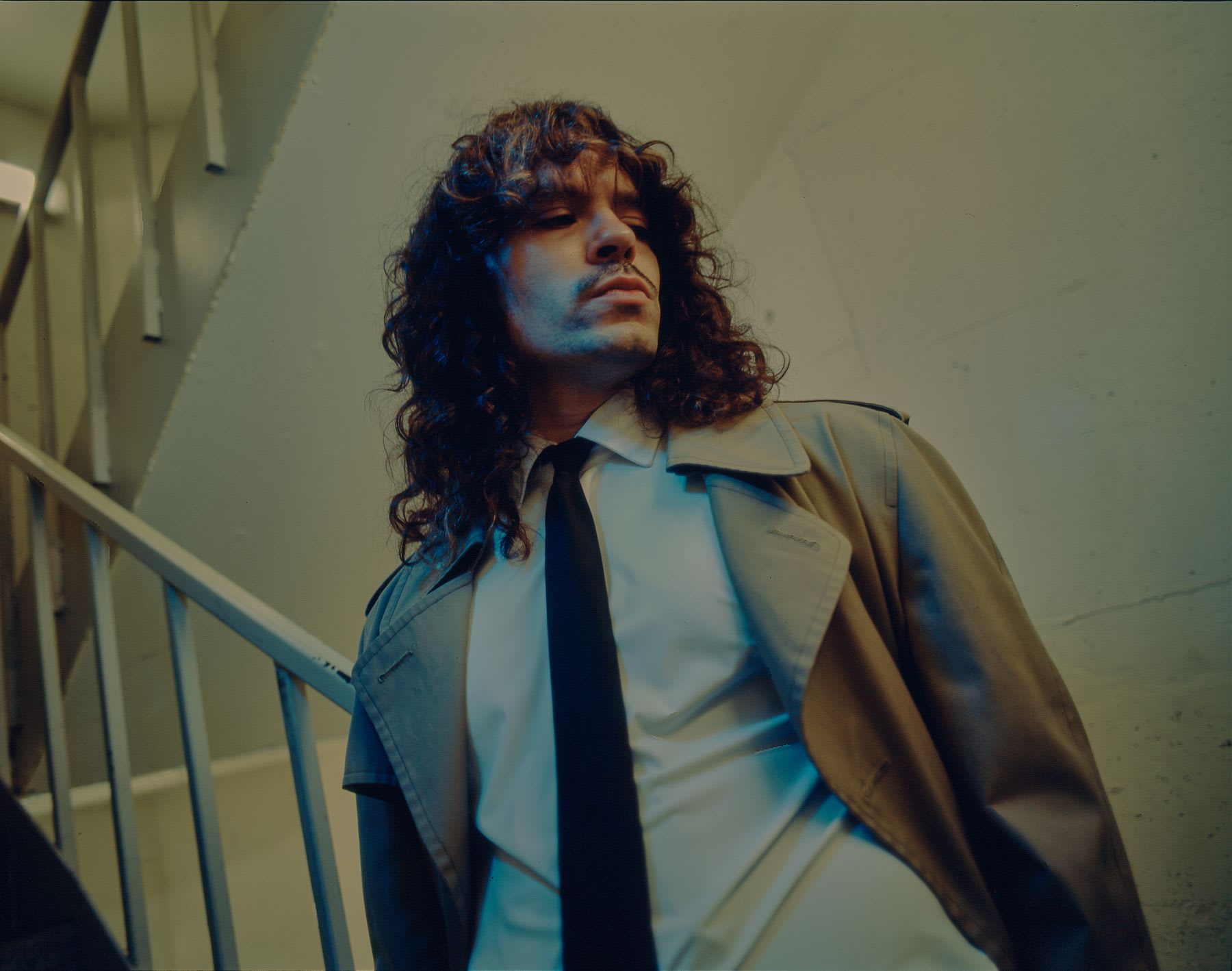 A man with long curly hair wearing a trench coat looks down with a pensive expression while standing in a dimly lit stairwell.