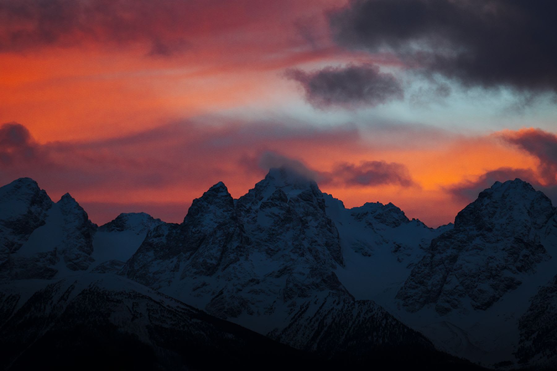 Sunset behind snow-covered mountain peaks with clouds illuminated by warm hues of orange and red