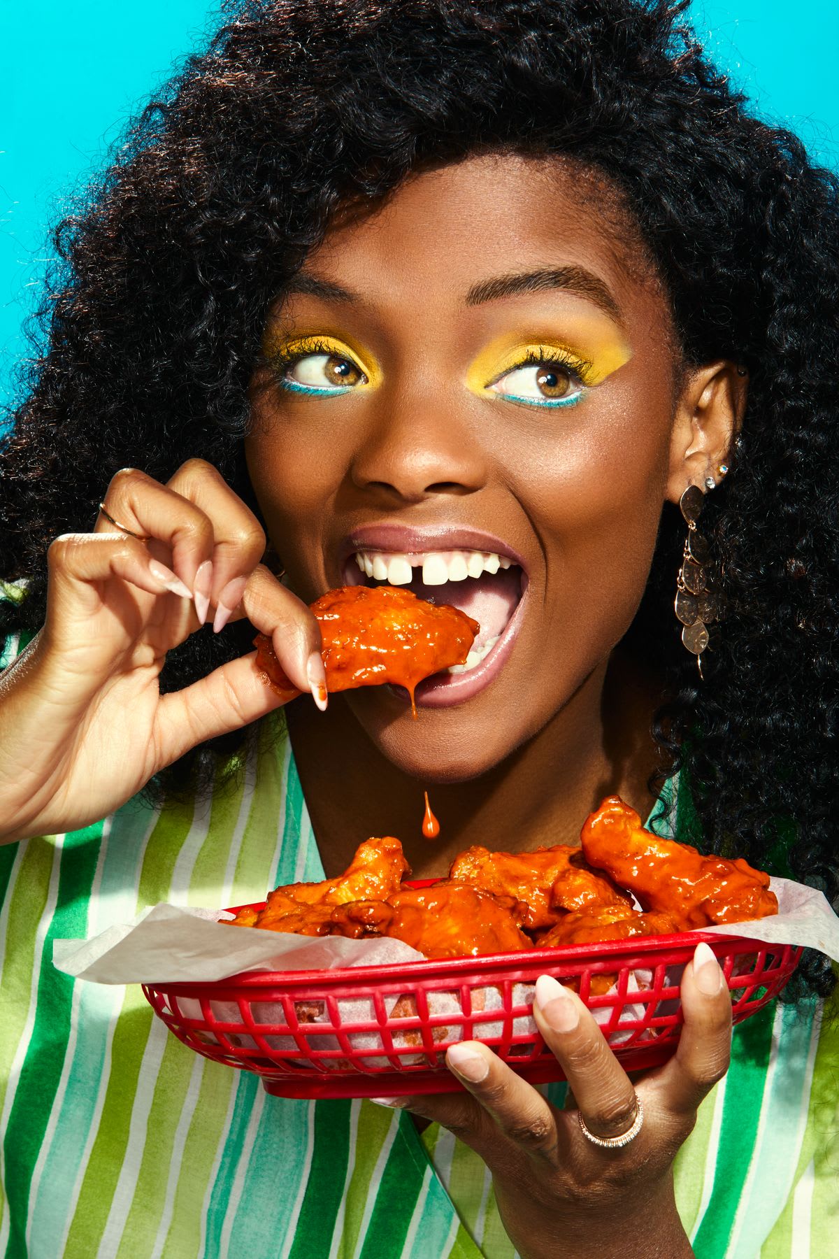 a woman with dark curly hair and striking yellow and blue eye makeup eagerly biting into a saucy chicken wing held above a red basket filled with more wings.