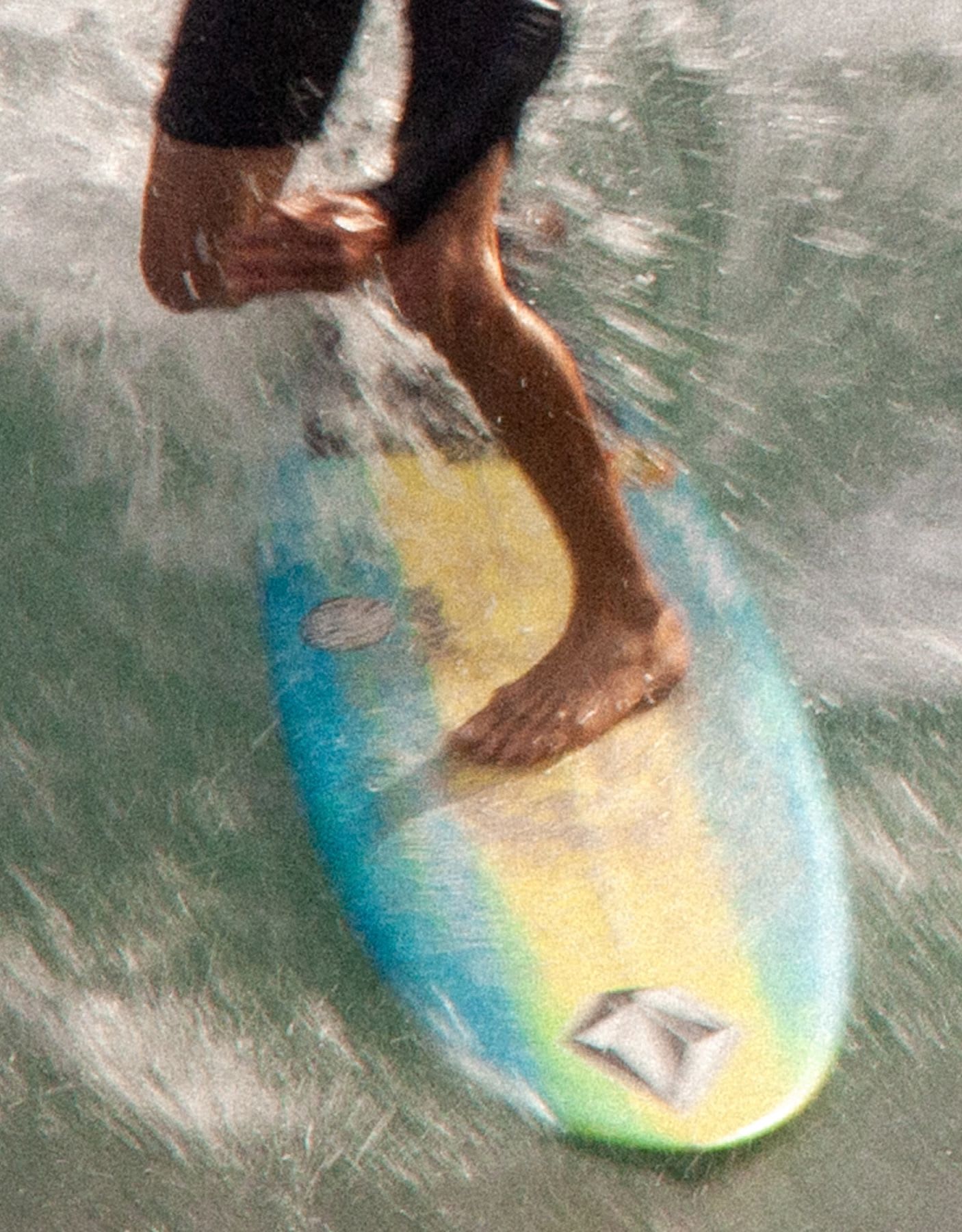 A close-up, grainy photograph captures a surfer's feet balancing on a colorful board while riding a wave.