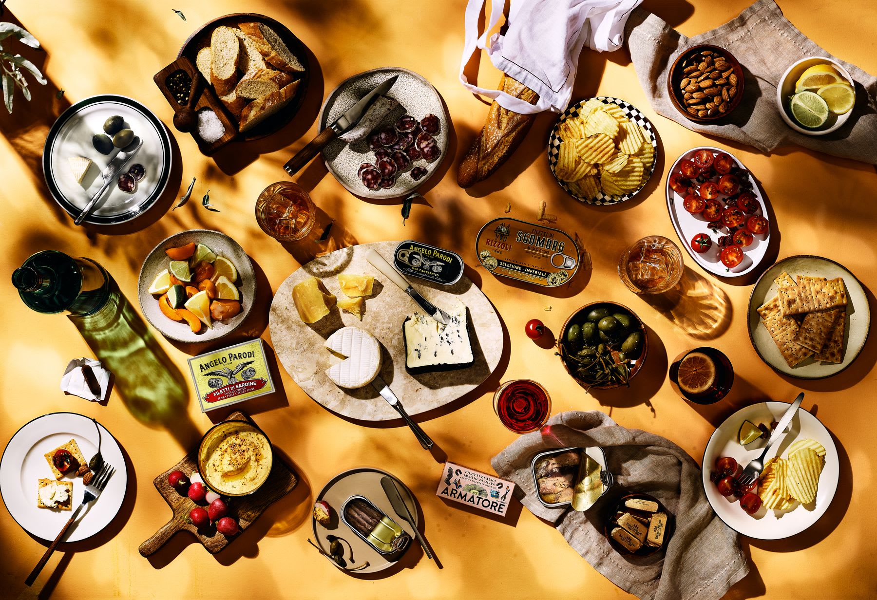 An outdoor snack table with various foods such as cheeses, olives, bread, drinks, arranged on plates and boards on a yellow surface.
