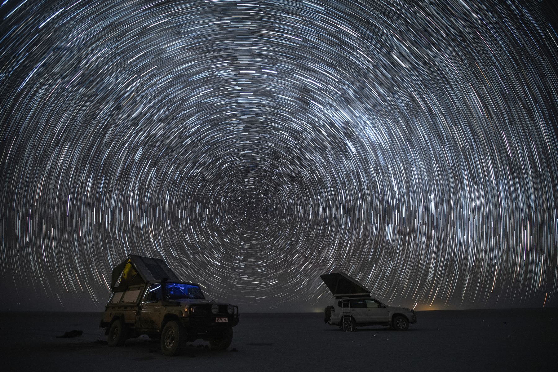 Two vehicles under a mesmerizing star trail sky, indicating the rotation of the Earth as captured in a long-exposure night photograph.