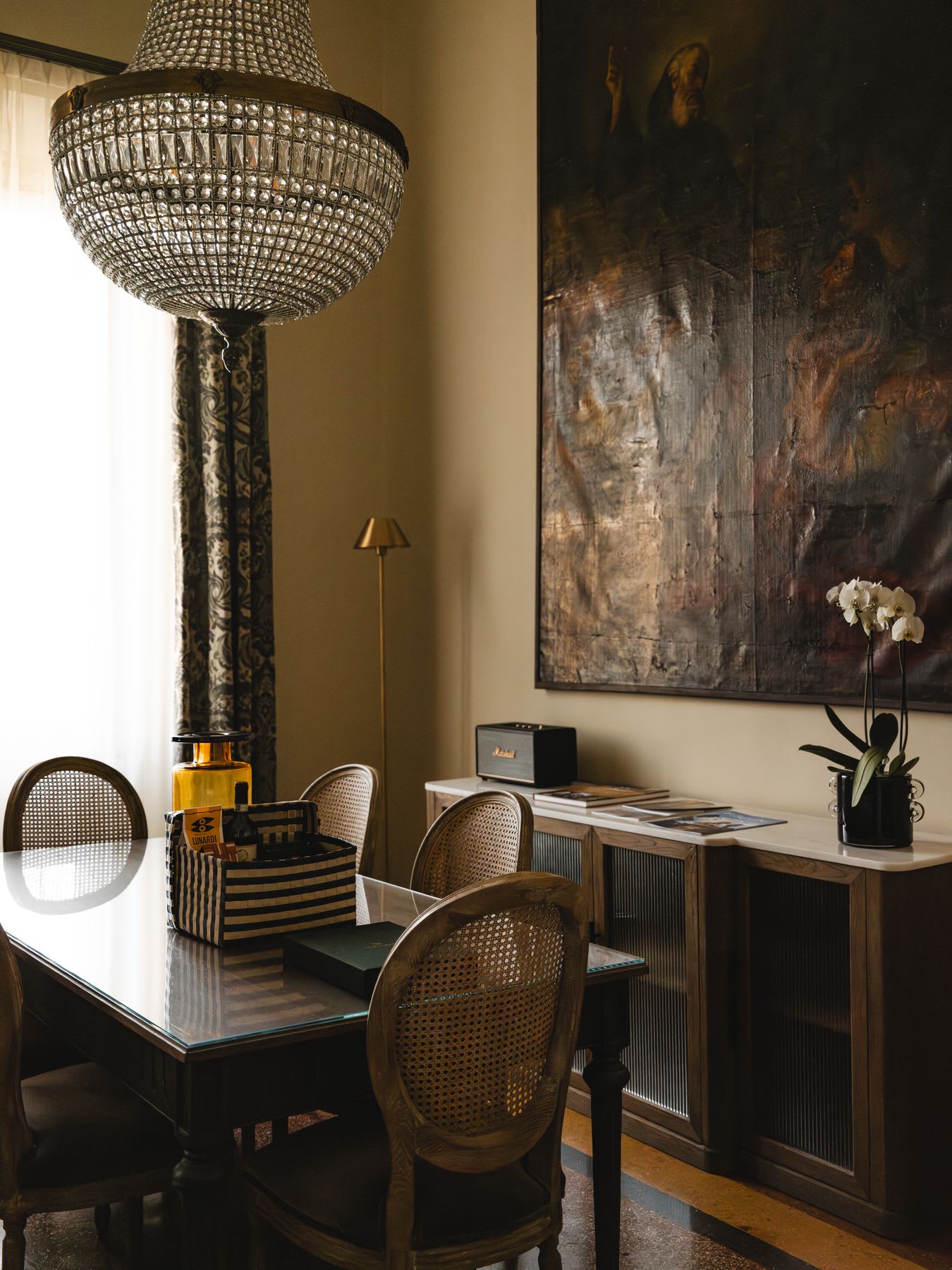 A large crystal chandelier hangs over a dining table with cane chairs in a room decorated with a sideboard and large, dark artwork.