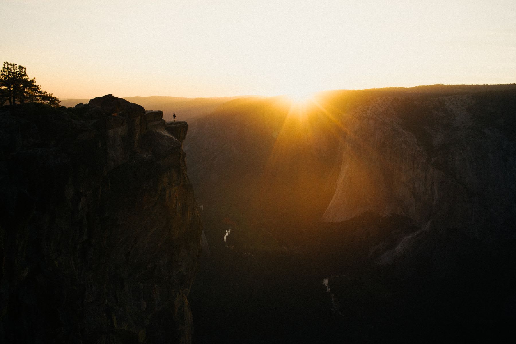 A lone person stands silhouetted on the edge of a massive cliff overlooking a deep valley during a golden sunset.