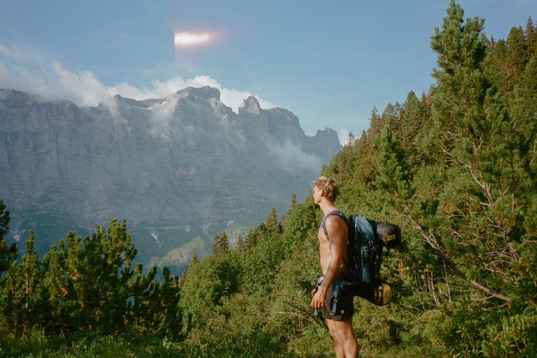 A young man pauses to look at some nearby mountains while backpacking near a forest.