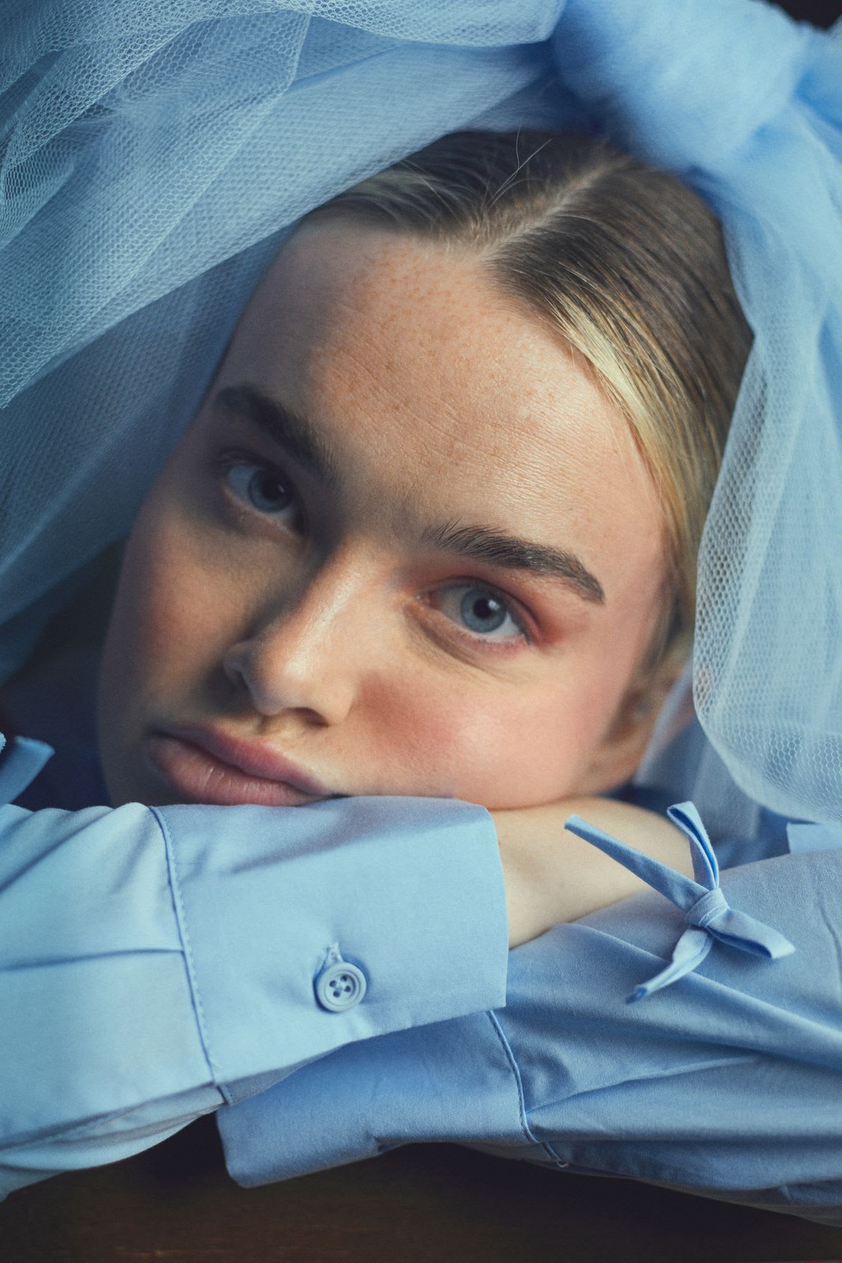 A woman with blue eyes rests her head on her arms, wearing a light blue shirt adorned with small bows and a large tulle bow on her head.