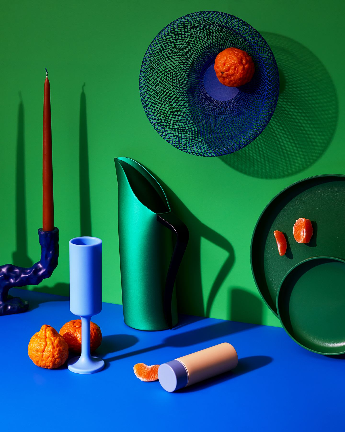 A simple still life with a green pitcher, a bowl of oranges, and two unlit candles on a blue wooden table.