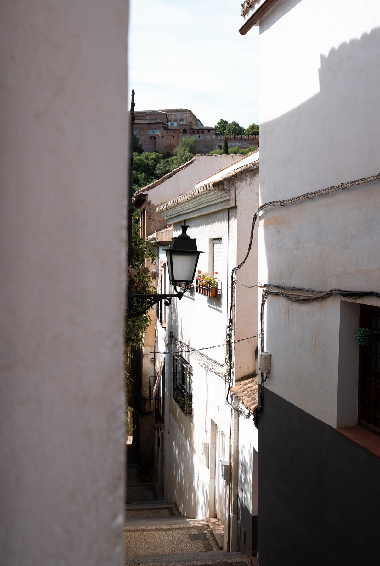 Sunny narrow street lined with white buildings and a vintage lamp post, with a view of a distant historic building on a hill.