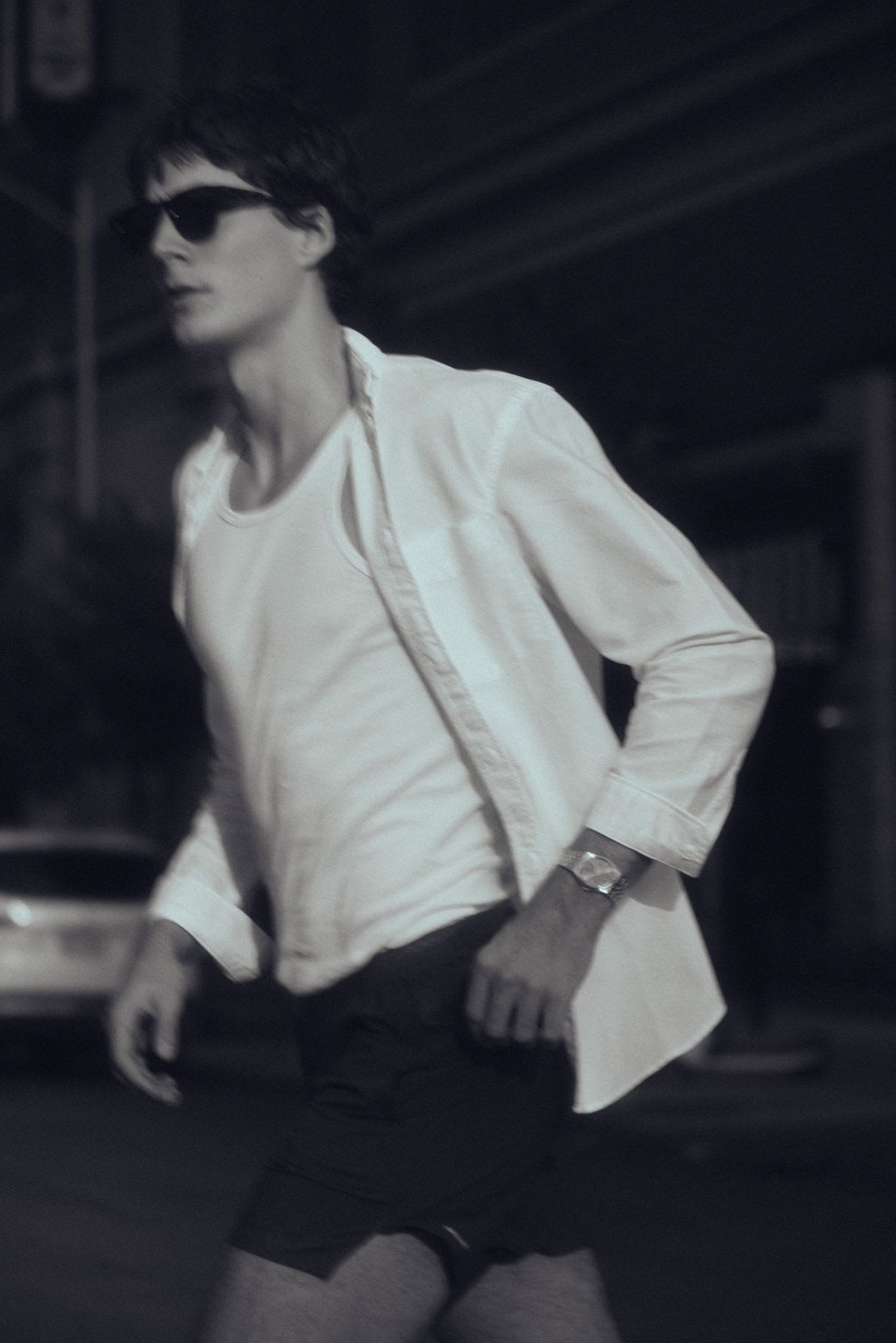 A stylish man in sunglasses and an unbuttoned white shirt walks down a street at night in a candid, monochrome photograph.