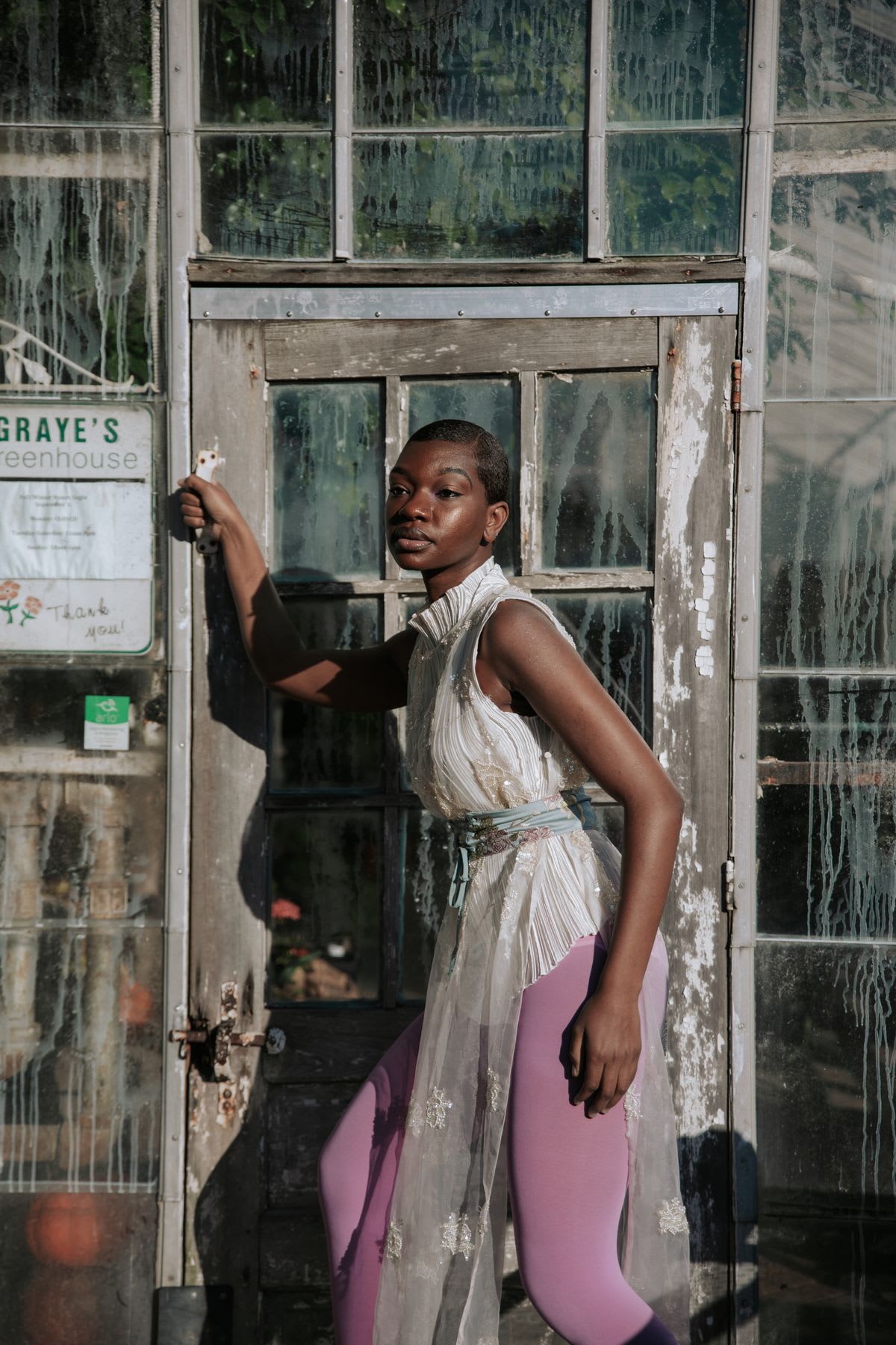 Woman next to a glass door in a greenhouse wearing a white dress and pink pants