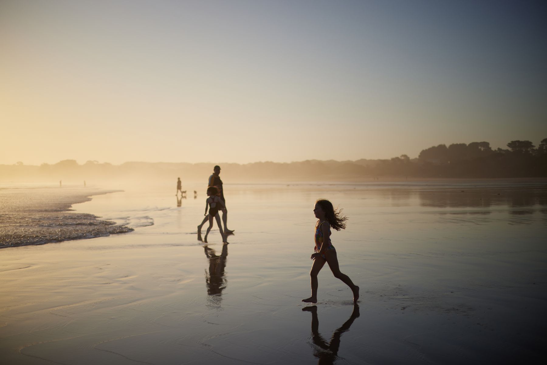 Kids run around in shallow water at the beach with their father