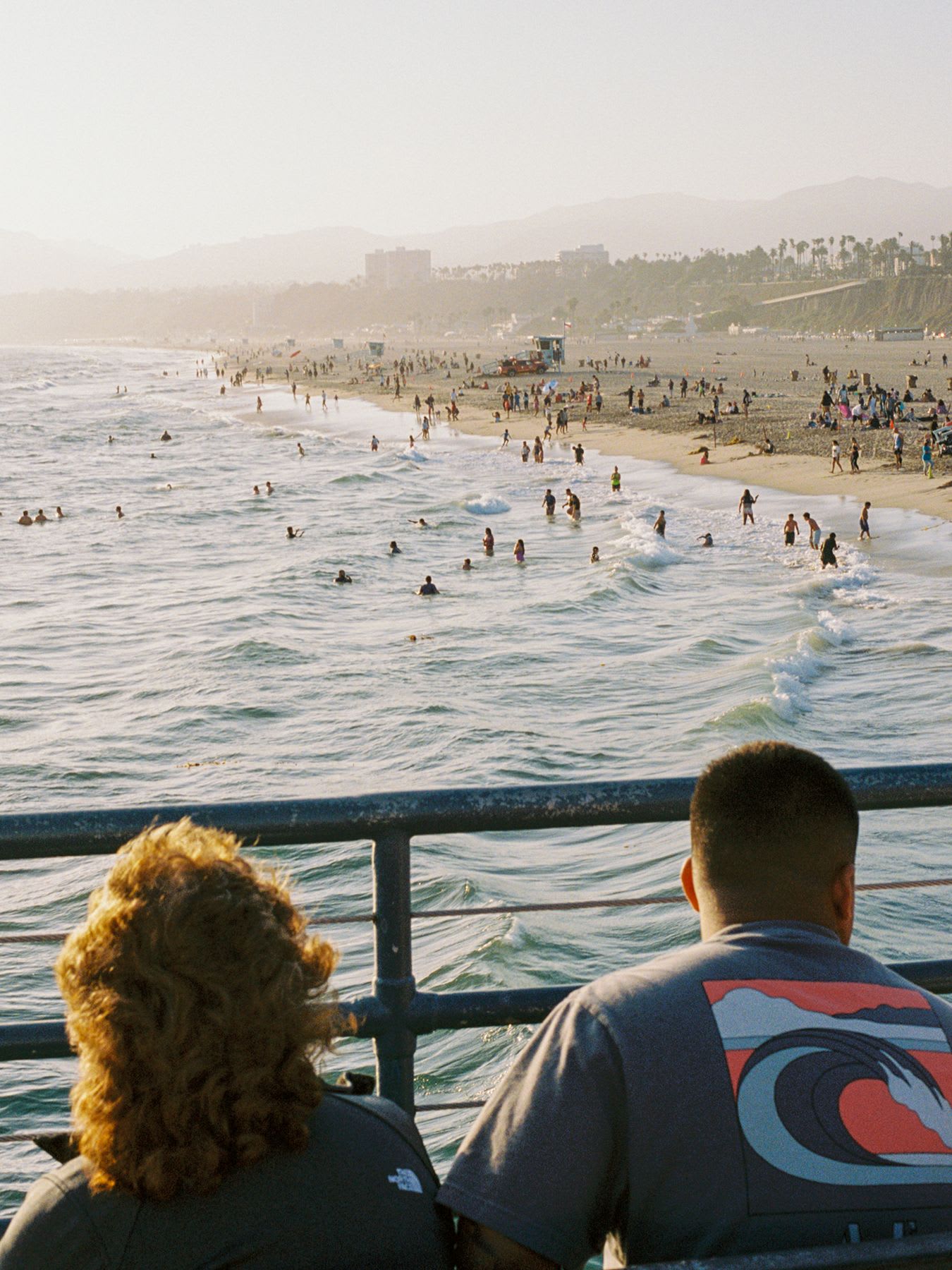 A man and a woman watch from a railing as crowds of people swim and relax on a busy beach on a hazy summer day.
