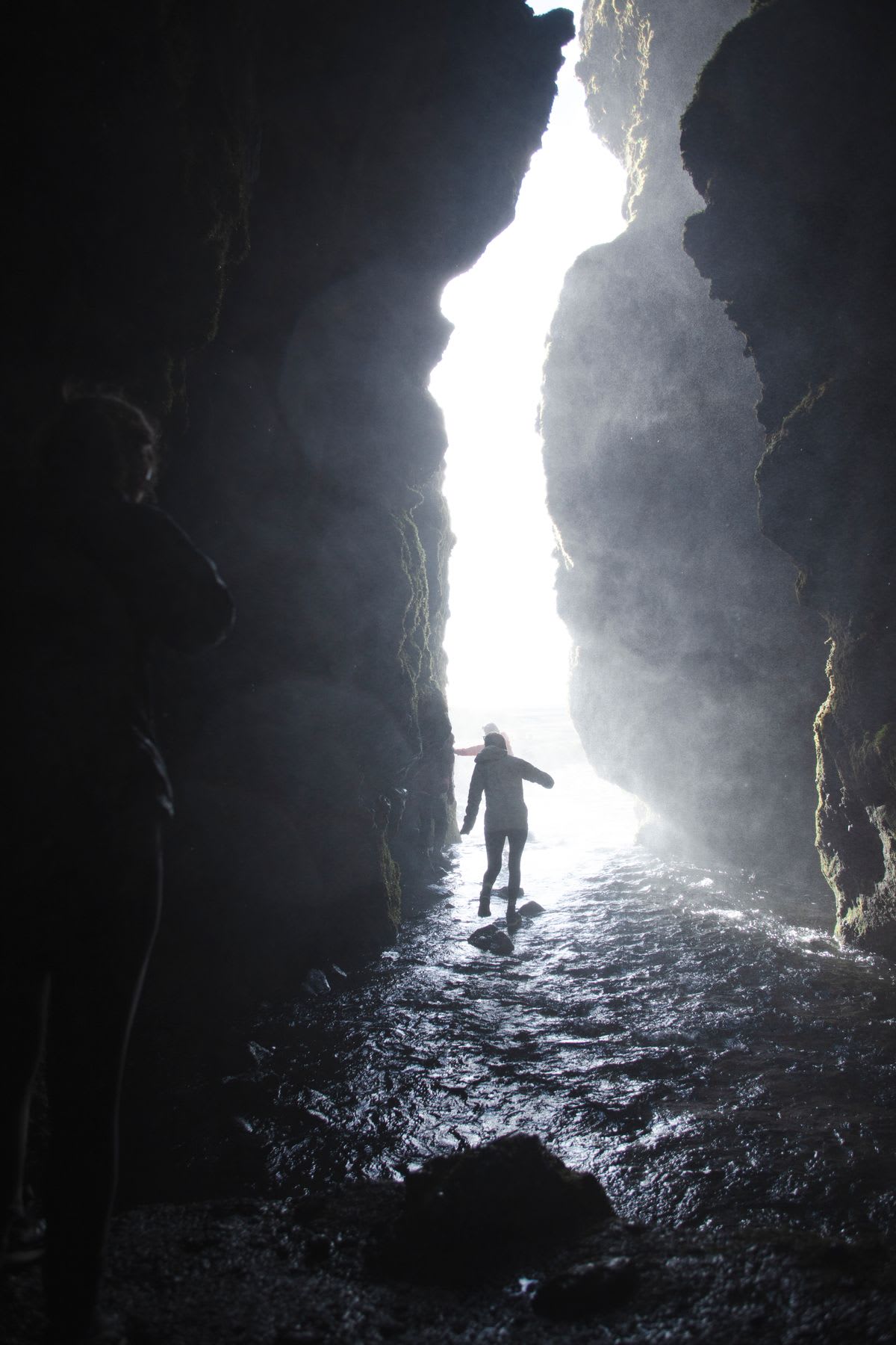 Two individuals emerge from a rocky cave and walk into the ocean during the daytime.