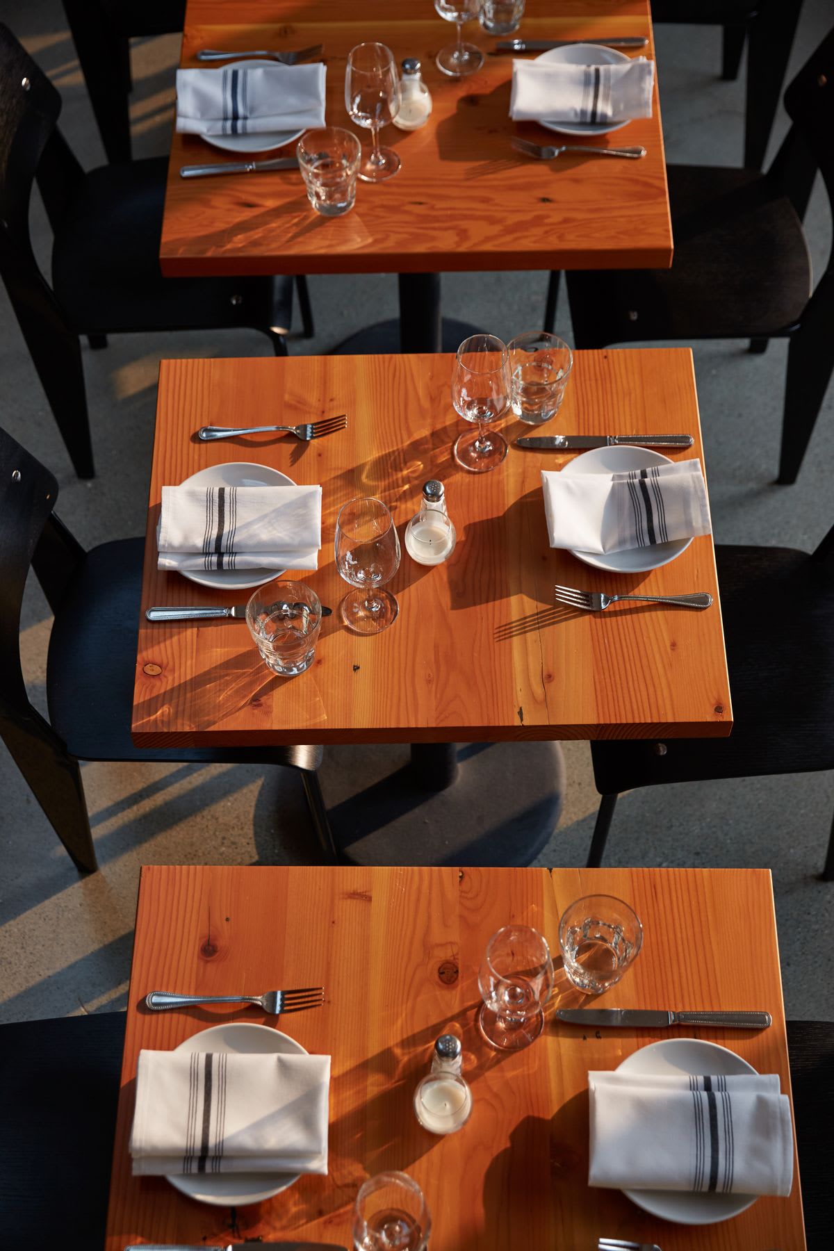Wooden restaurant tables are neatly set with white plates, striped napkins, silverware, wine glasses, and salt shakers, surrounded by black chairs in warm sunlight casting long shadows.