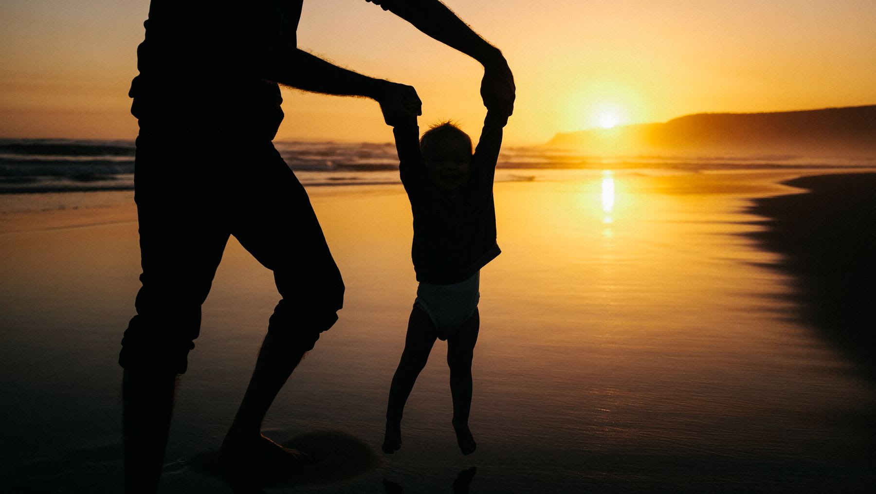 A father plays with his young toddler on the beach at sunset.