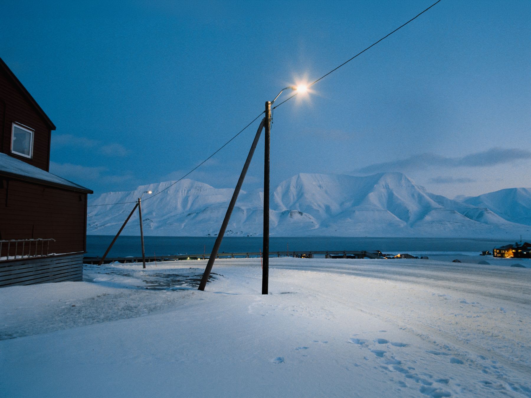 A street lamp illuminates a snow-covered road in a remote coastal village against a backdrop of towering, snowy mountains during the blue hour.