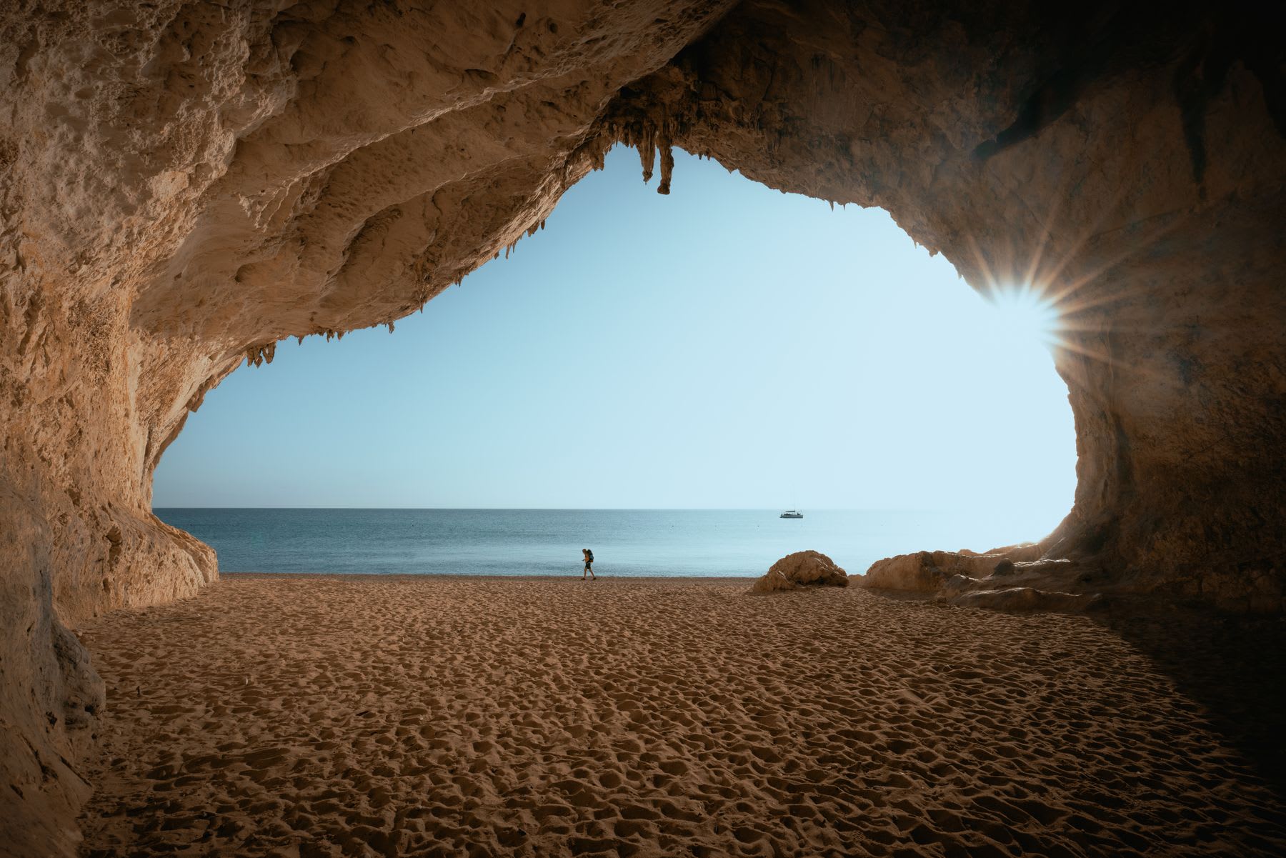 A backpacker walks along the ocean shore beside a deep cave on a sunny day.