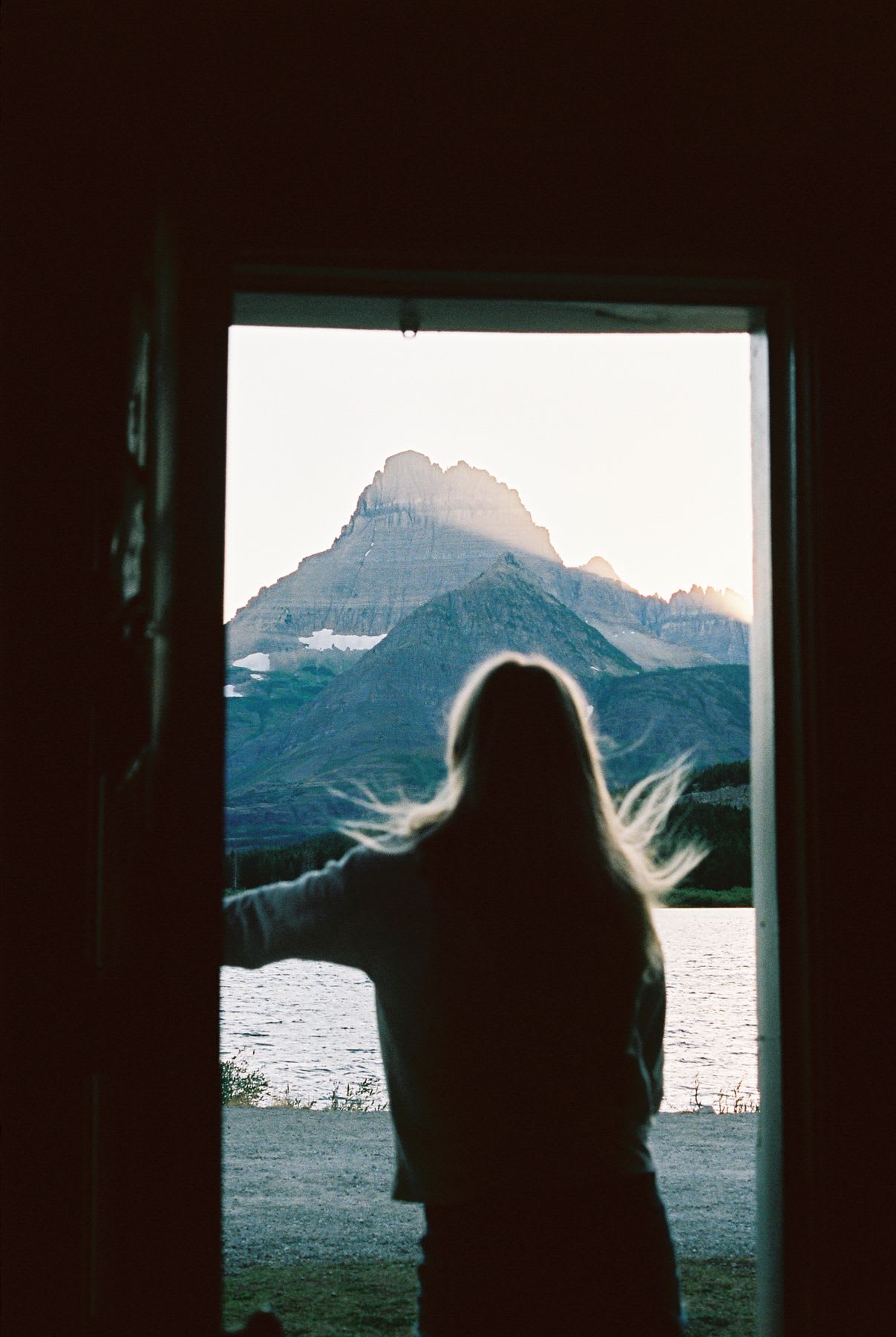 A silhouetted woman with wind-blown blonde hair stands in a doorway looking out at a majestic mountain peak over a lake at sunrise.