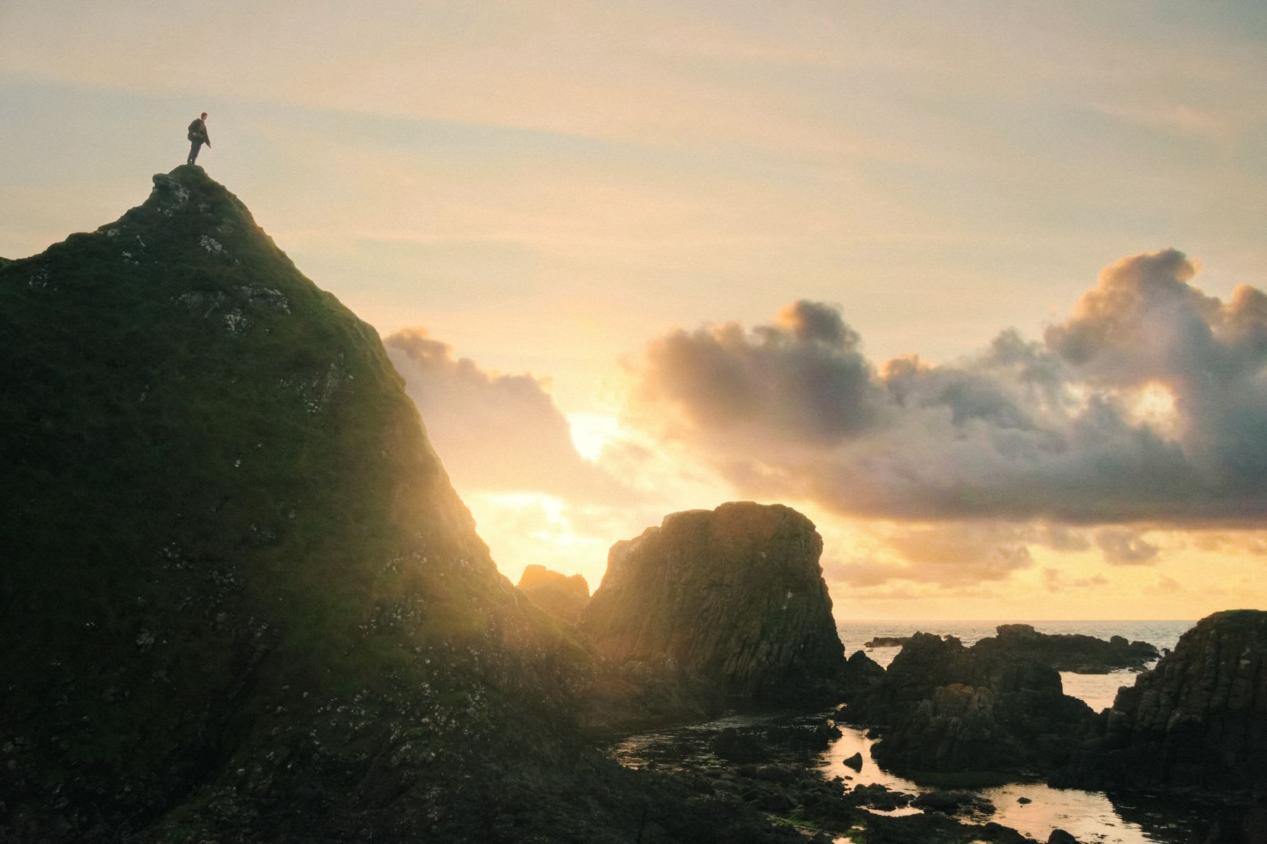 A hiker stands on top of a mountain on a rocky coastline at sunrise.