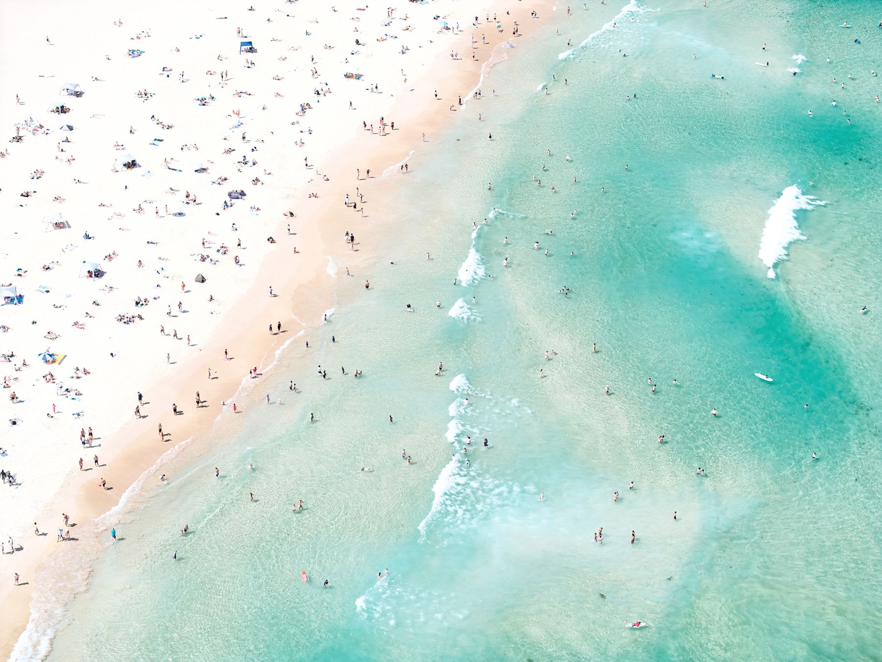 A beach full of people enjoying the sun and the sea, many of them in the water and others relaxing on the sand.