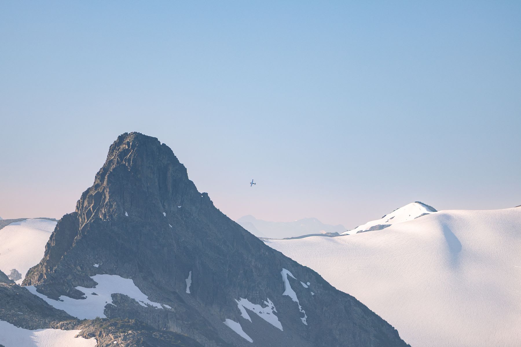 A small white airplane flies past a jagged, rocky mountain peak surrounded by snowy glaciers under a clear blue sky.