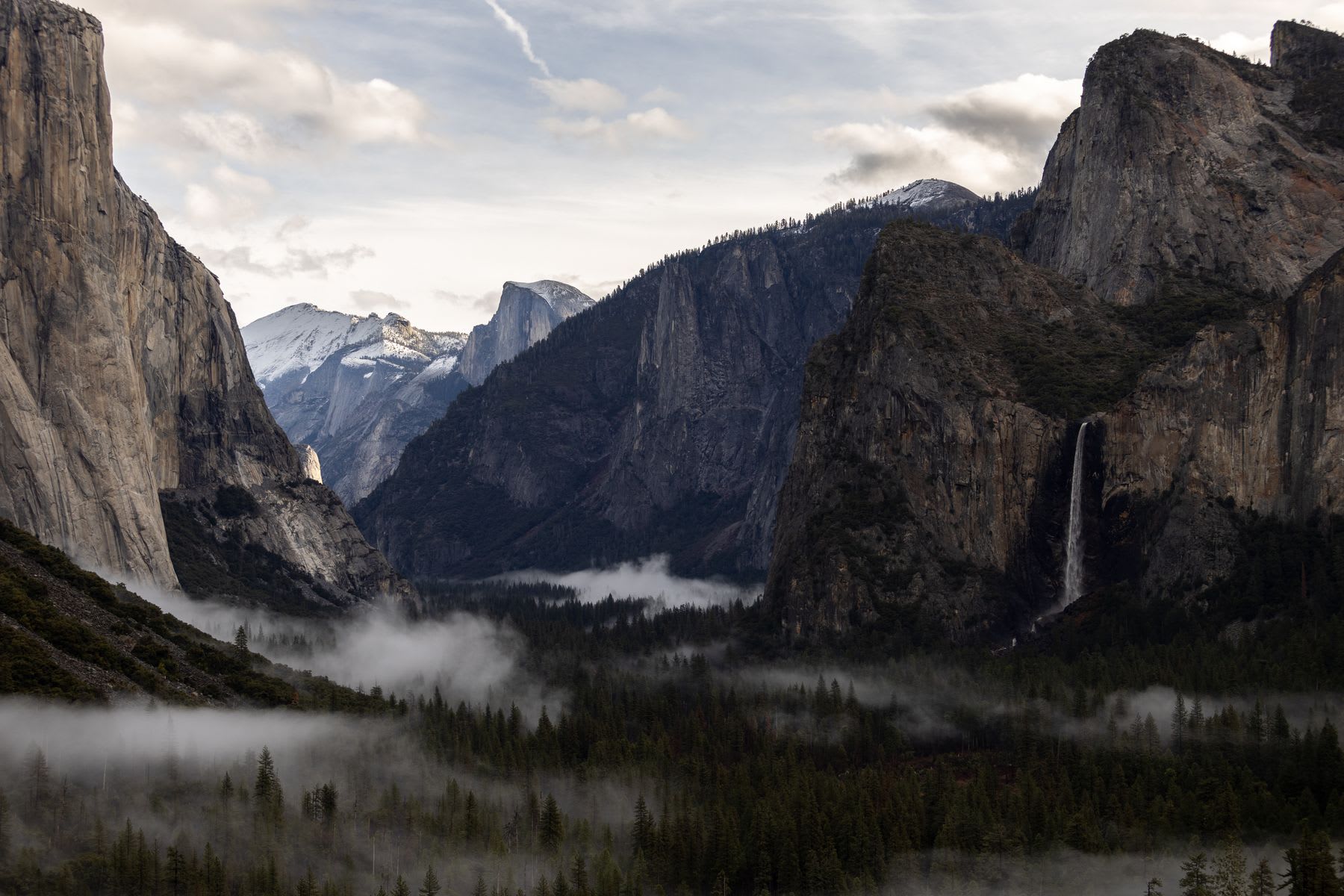 An iconic view of Yosemite Valley where low-hanging fog drifts through the forest, framed by granite cliffs and a cascading waterfall.