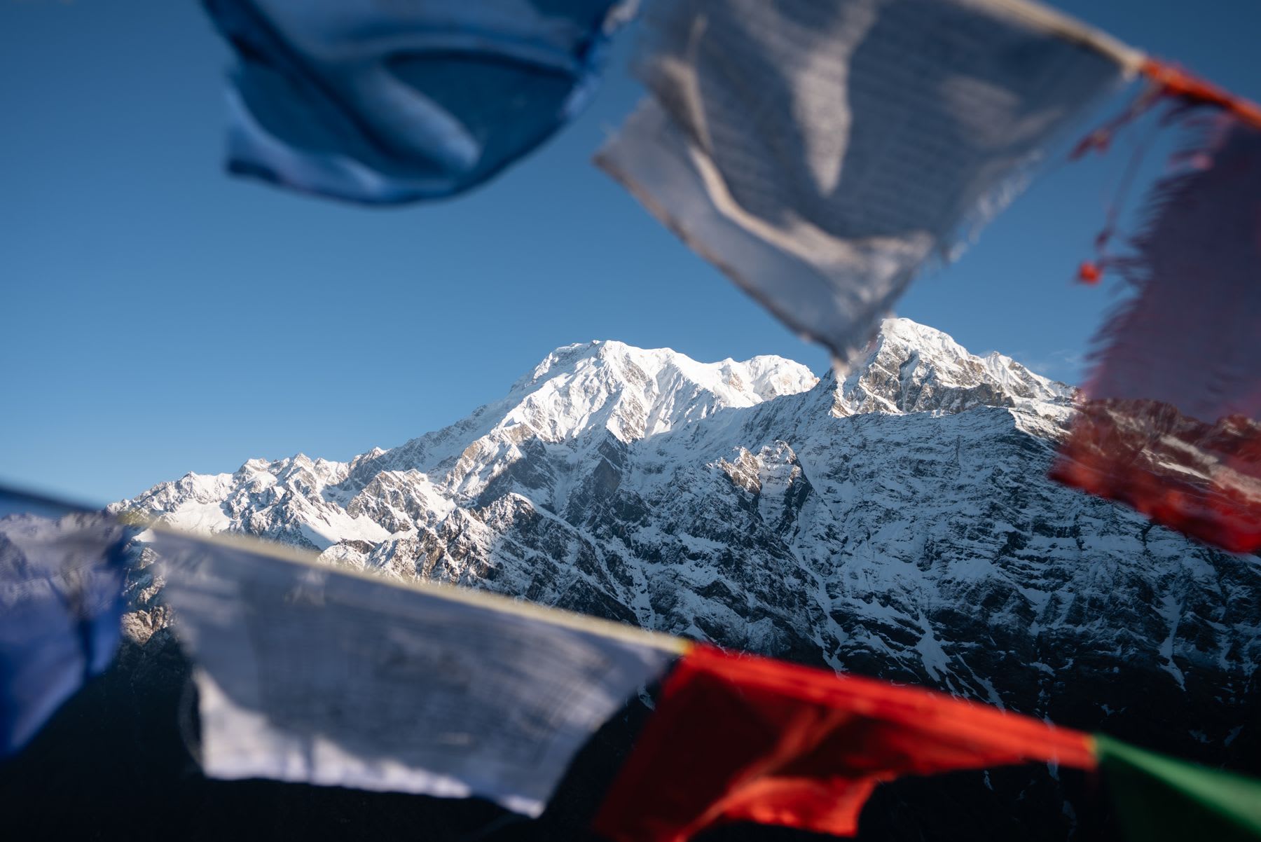 Colorful flags flutter in the foreground before an imposing range of snow-capped mountains under a blue sky.